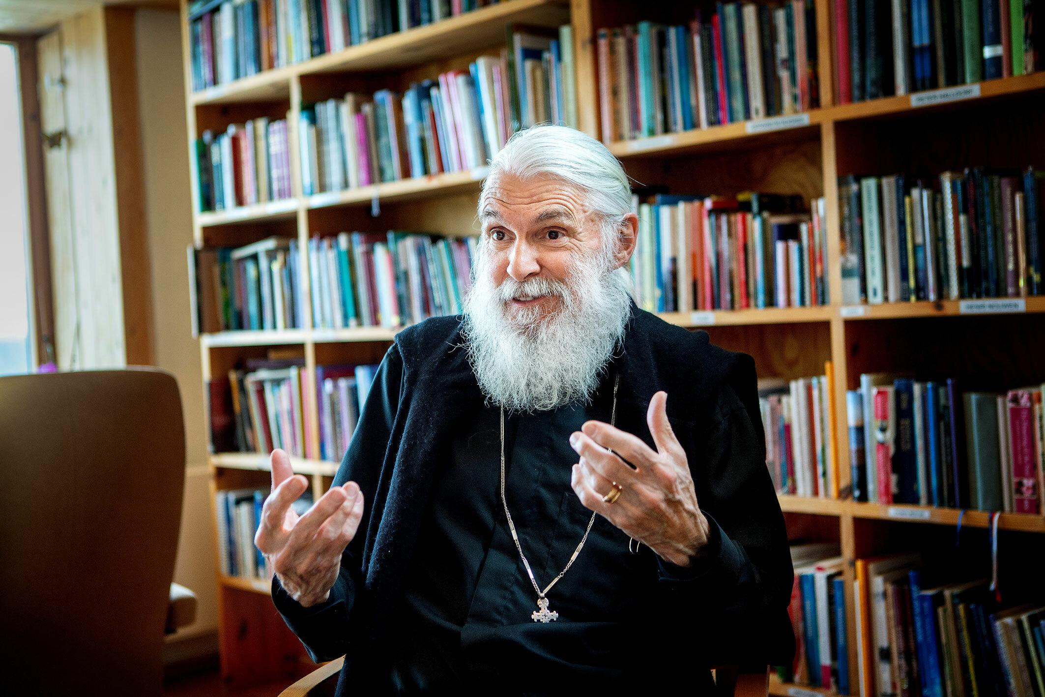 Foto: Mann med gråhvitt hår og skjegg gestikulerer med hendene mens han prater. Han sitter foran en hel vegg med bøker, iført svarte klær og et ortodoks kors.