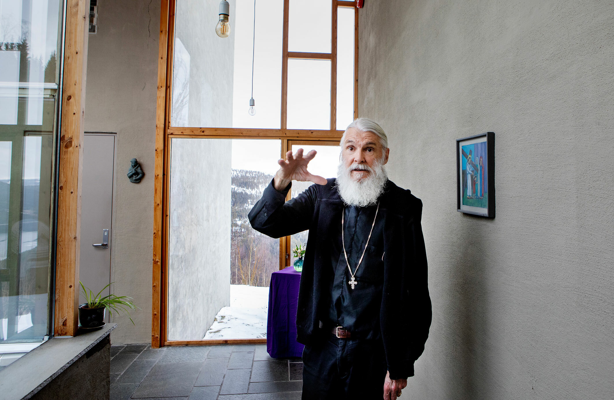 Foto: Mann i svarte klær, kors og mye hår og skjegg står foran et vindu som går fra gulv til tak.