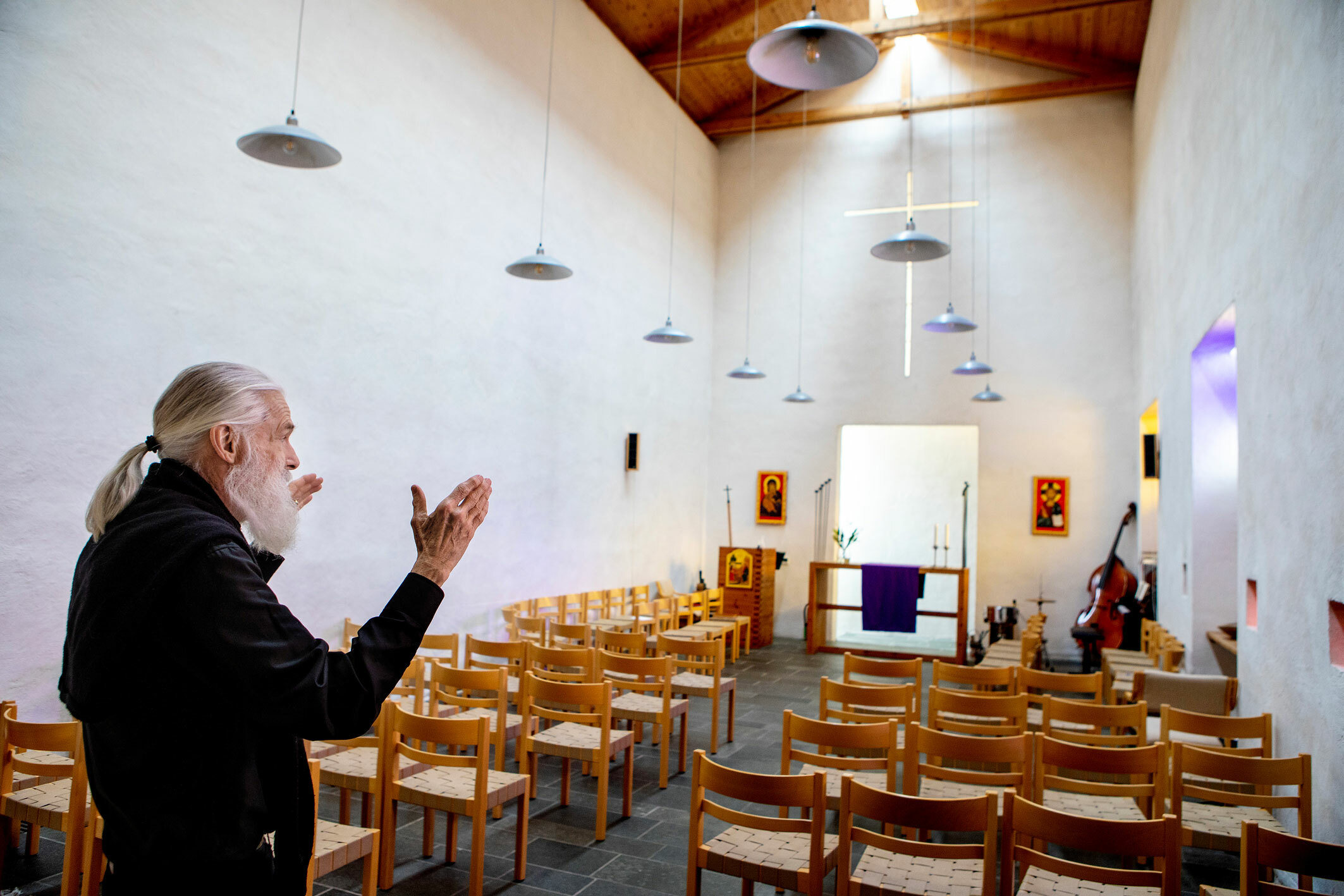 Foto: Mann med stort skjegg og grått hår i hestehale står med ryggen mot kamera. Han har svarte klær og står og ser innover i et lite hvitkalket kapell med enkle trestoler på rekker.