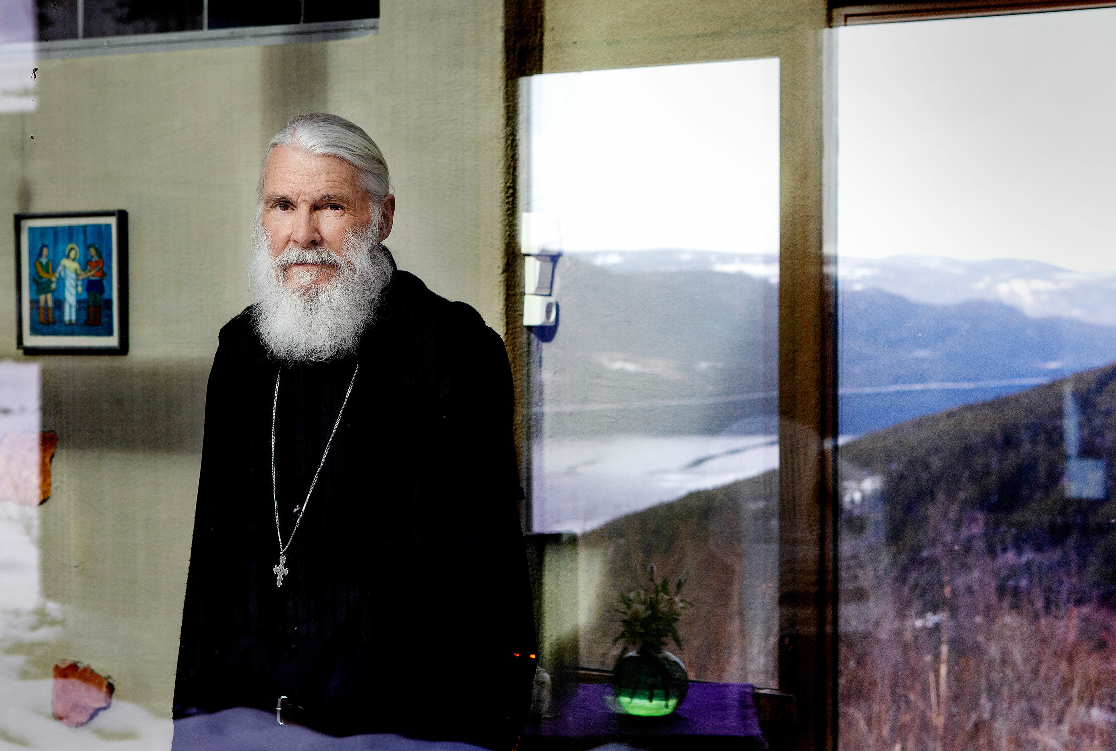 Foto: Mann med grått hår og skjegg ses gjennom et vindu. Han står oppreist foran et annet vindu med utsikt over fjell og fjord.