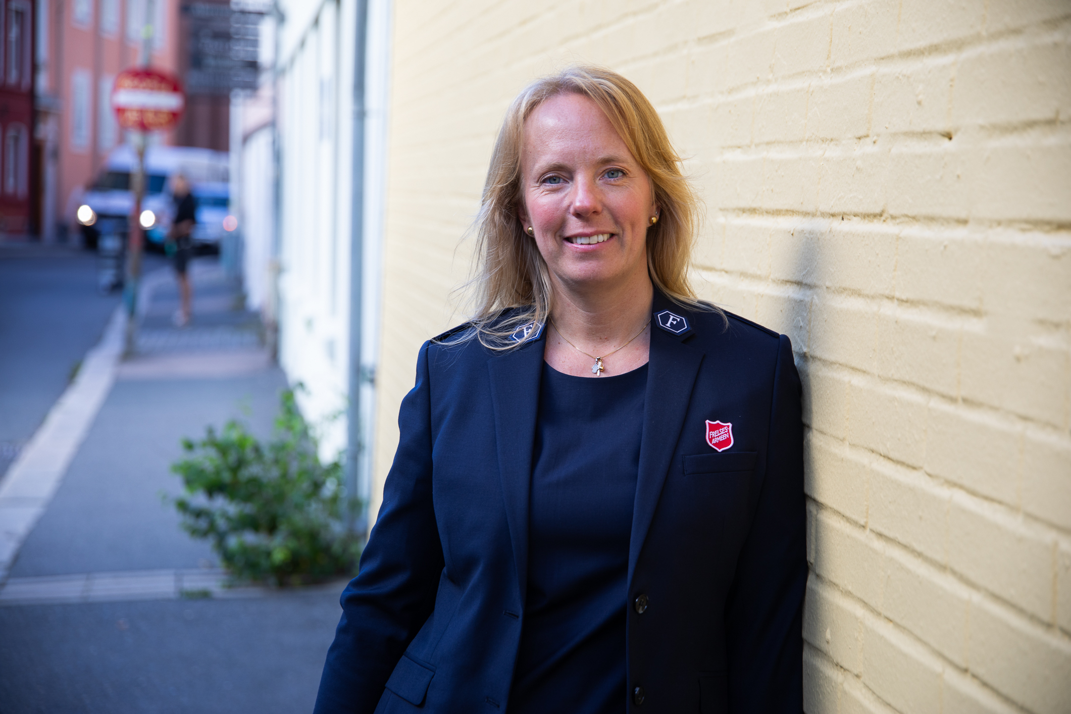 Kvinne i mørkeblå dressjakke og T-skjorte står lent inntil en gul murvegg ute i en gate. På jakkeslaget har hun Frelsesarmeens logo.