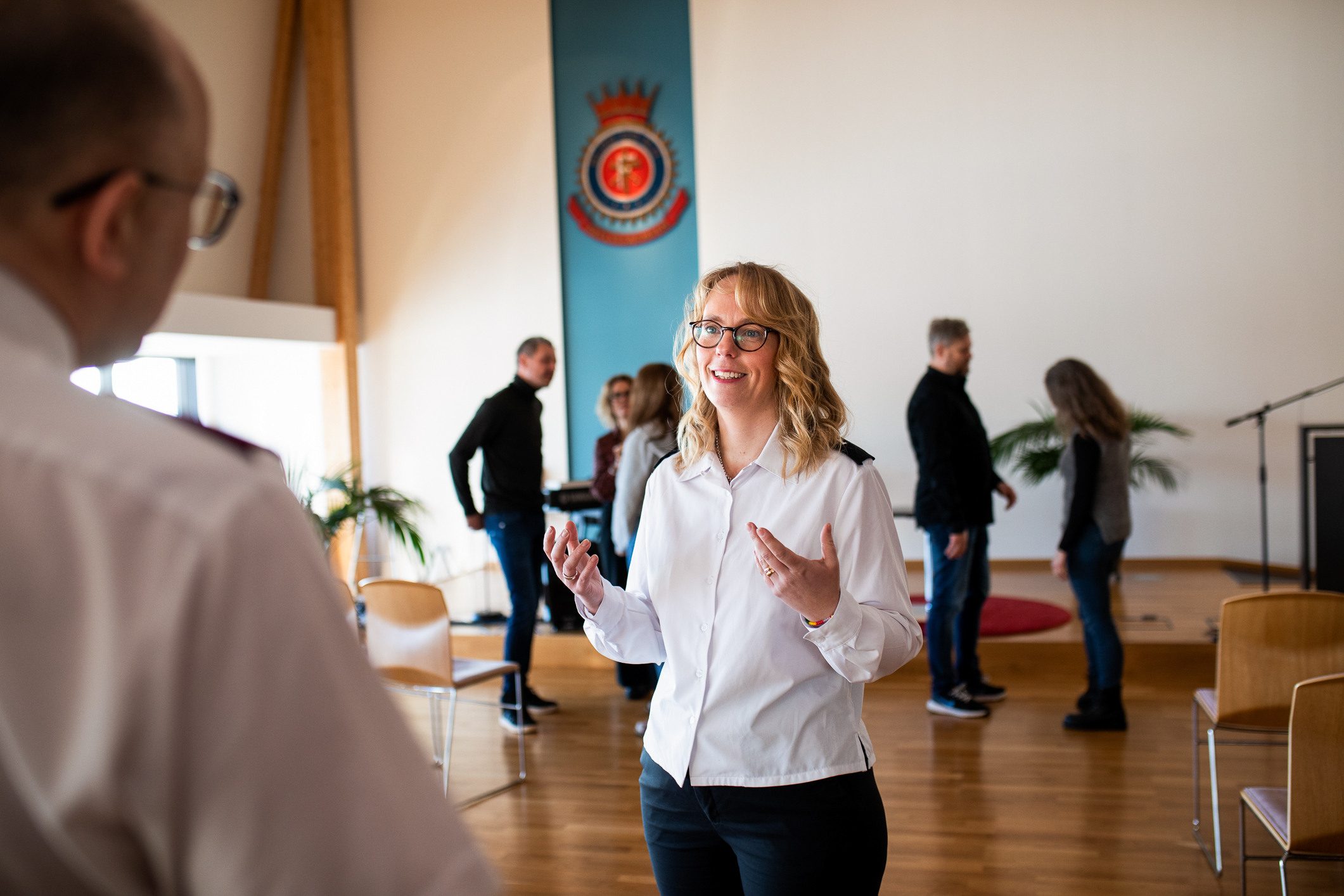 Kvinne står i en menighetssal. Folk i bakgrunnen og hun snakker med en mann vi bare ser ryggen til i venstre utkant av bildet.