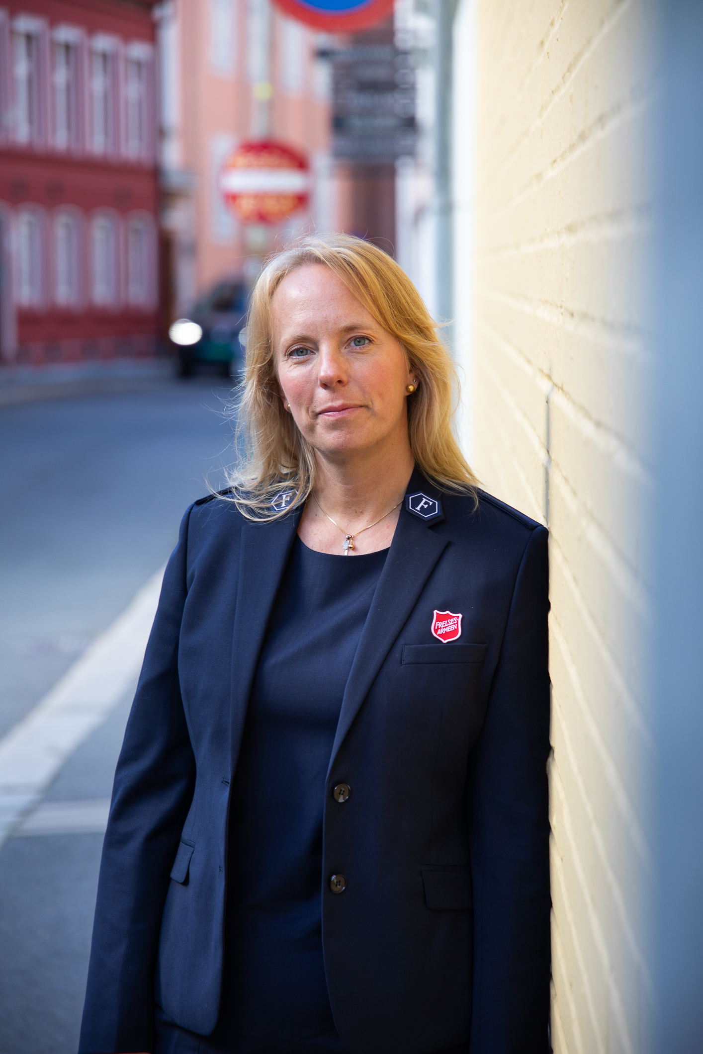 Kvinne i mørkeblå dressjakke og T-skjorte står lent inntil en gul murvegg ute i en gate. På jakkeslaget har hun Frelsesarmeens logo. Høydebilde.