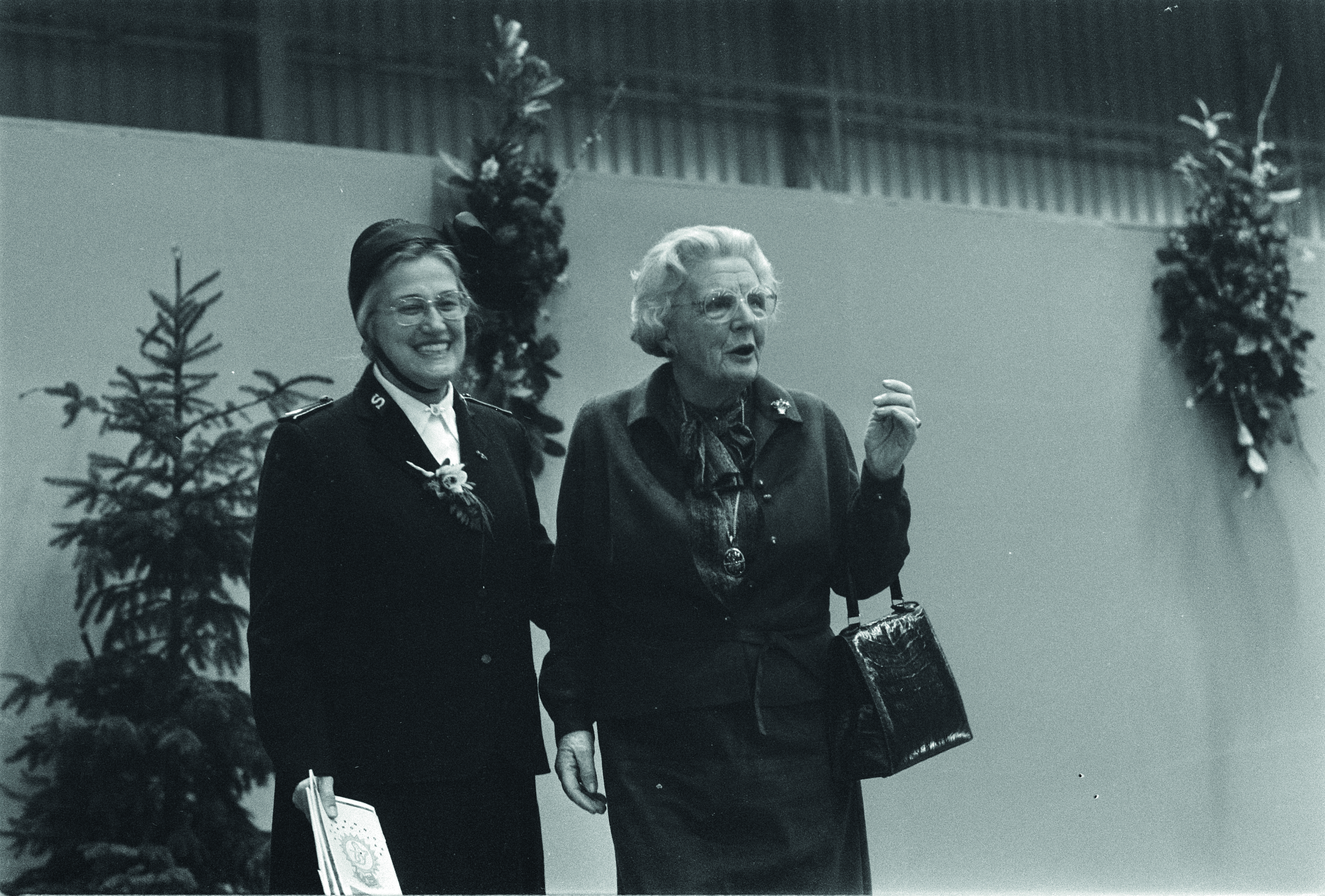 Et svart-hvitt bilde fra 60-tallet som viser to eldre kvinner. Til venstre står en kvinne med frelsesarméuniform og borett. Til høyre står en kvinne med drakt, veske, briller og hvitt hår lagt bakover.