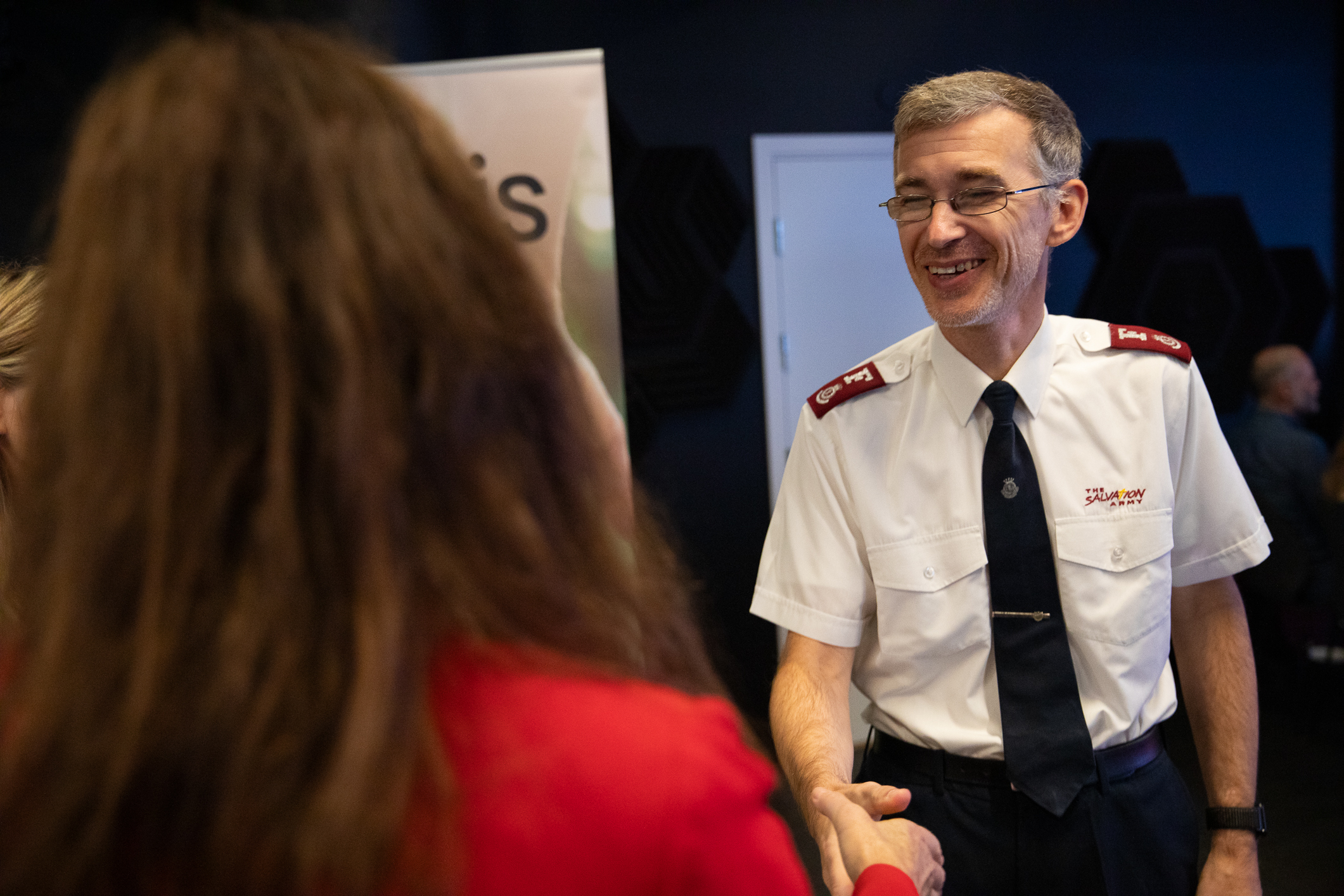 Foto: Mann med briller, i uniform fra Frelsesarmeen, hilser på en dame med svart, langt hår. Hun står med ryggen til.