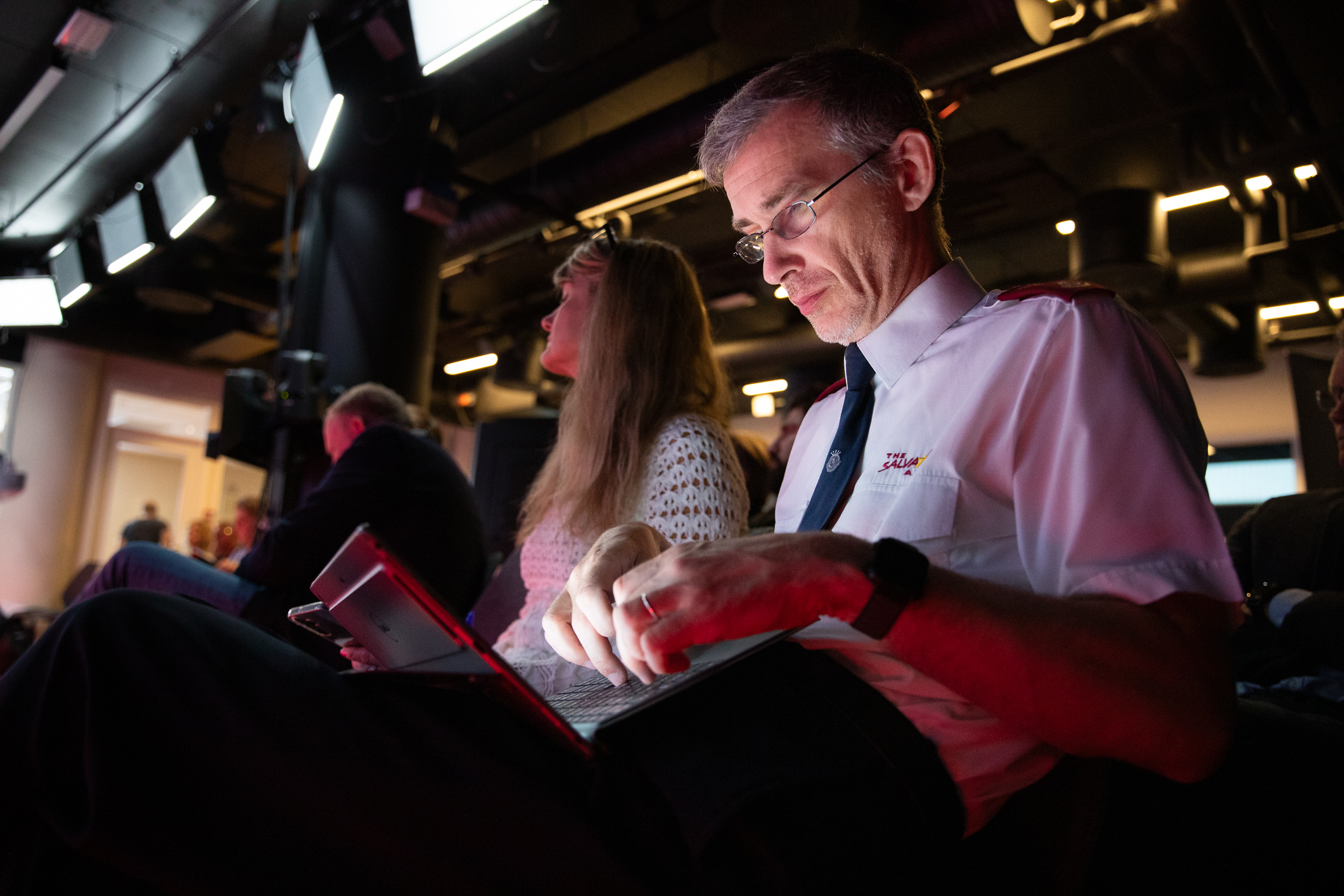 Foto: Mann med briller, i hvit uniformsskjorte, sitter og ser på PC-skjermen. På setes ved siden av ham i den mørke salen, sitter en blond kvinne.
