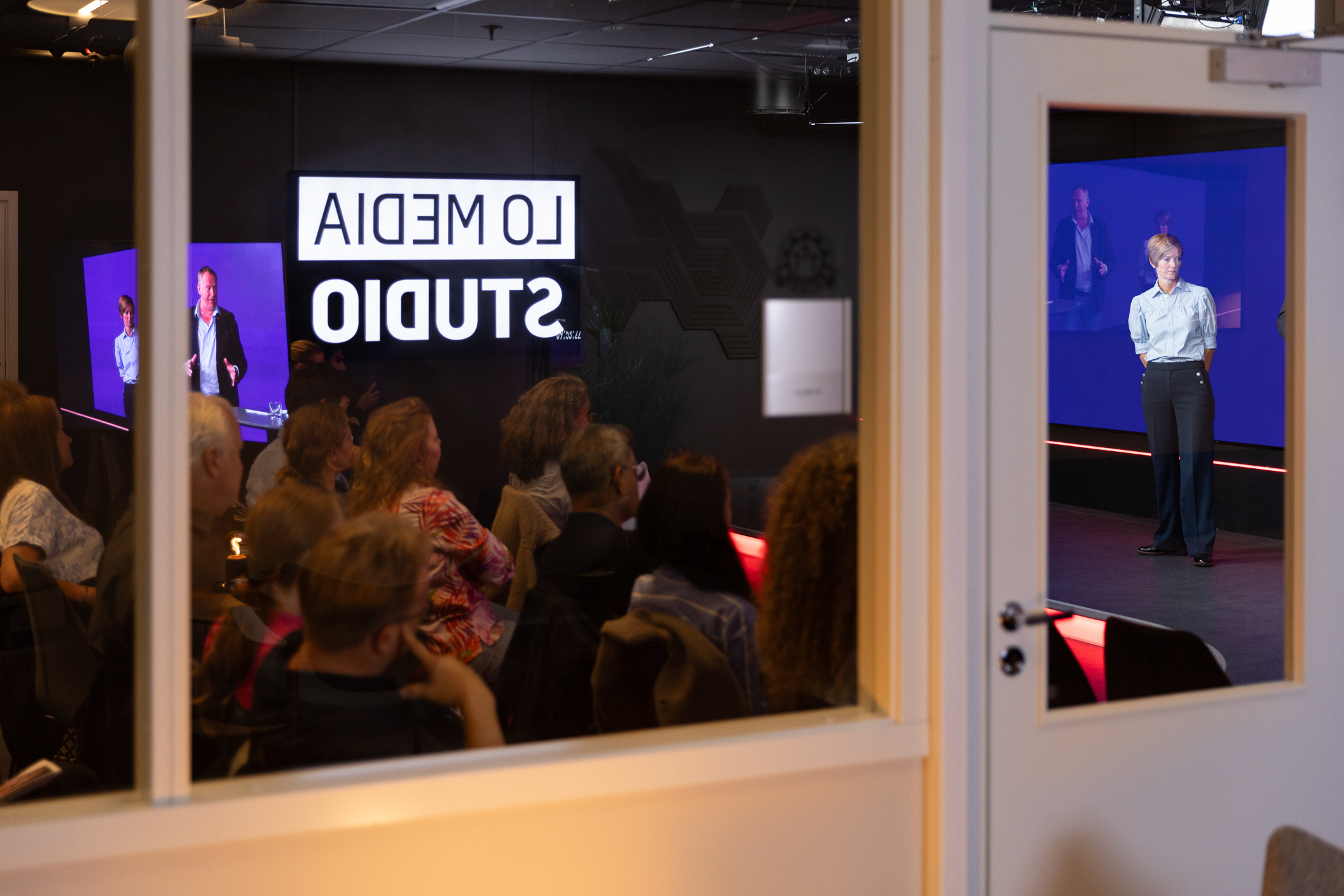 Foto: Vi ser inn gjennom vinduene til Lo Media Studio. Der står en mann og en kvinne på scenen. I den mørke salen sitter det folk og lytter.