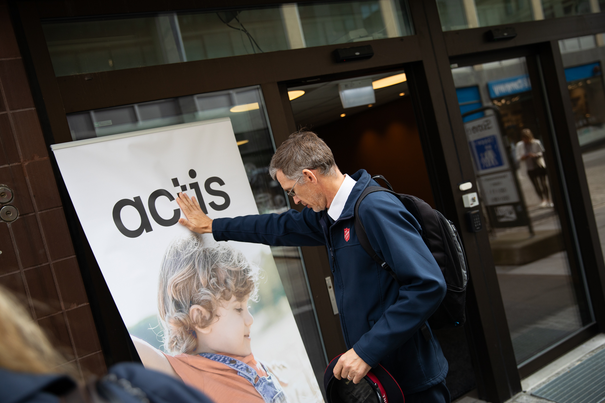 Foto: Mann med briller, i uniform fra Frelsesarmeen, holder hånden på en plakat det står Actis på.