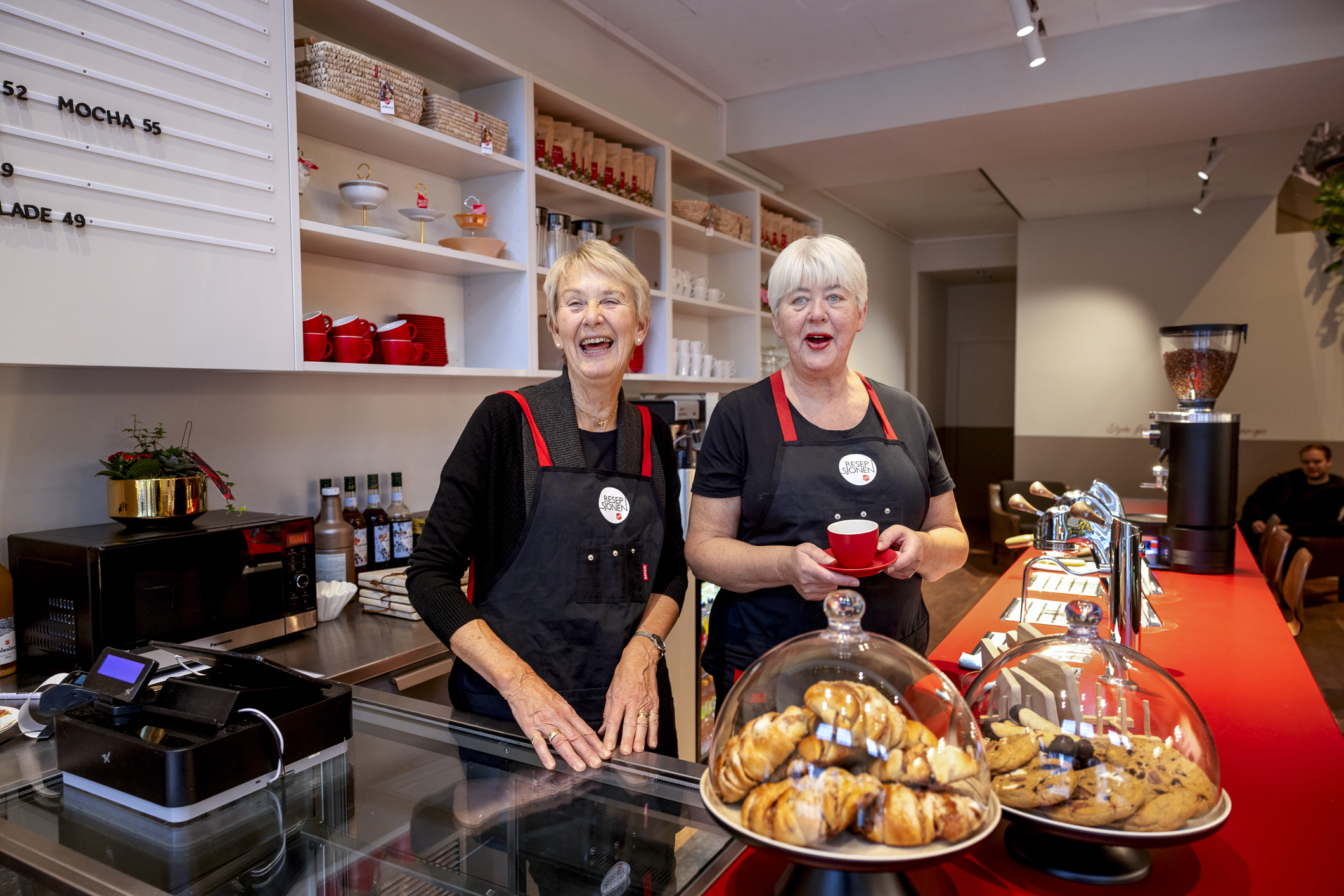 To gråhårede damer ler godt bak disken i kaffebaren. På disken står bakverk.