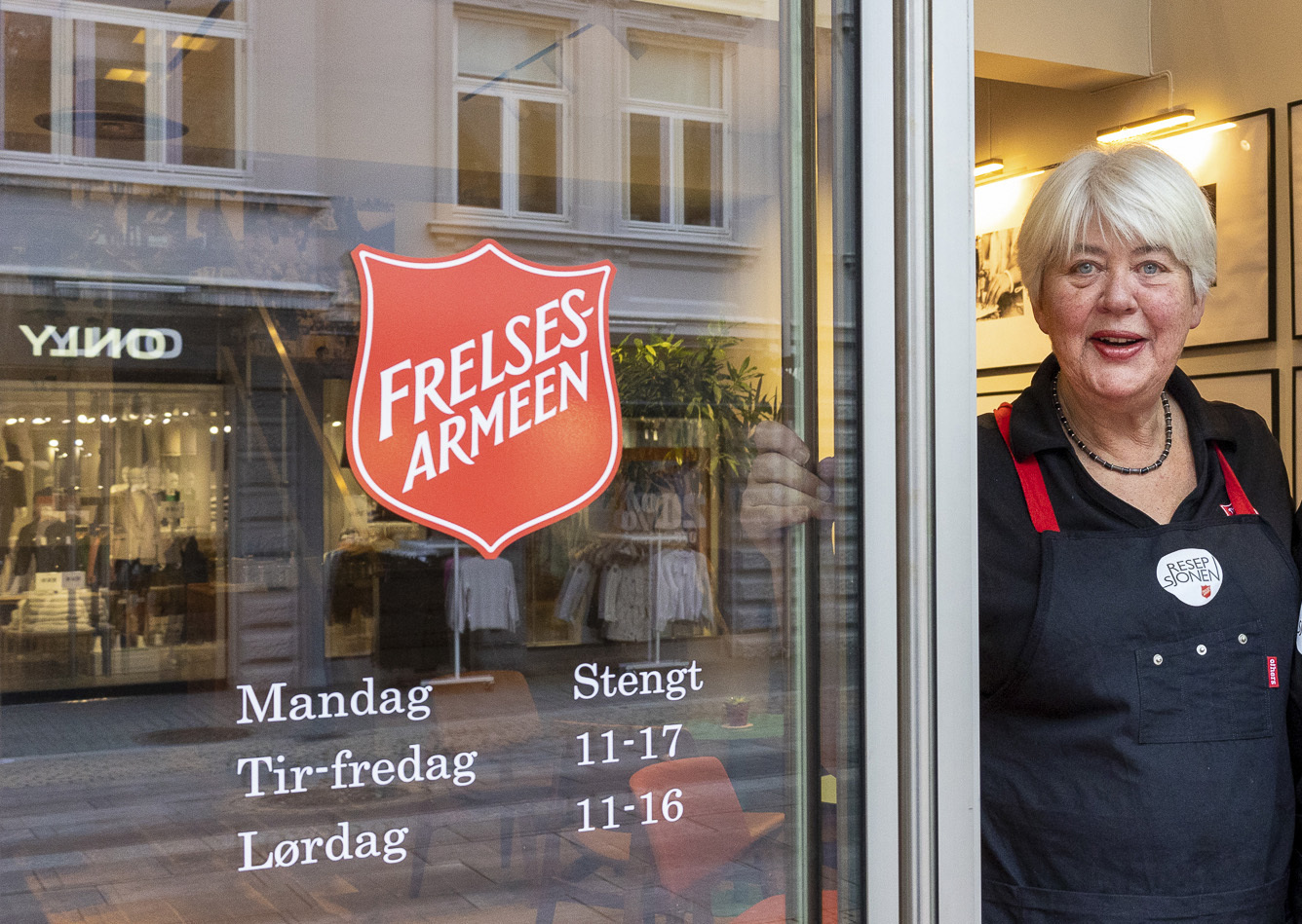 Kvinne i blå klær med forkle står i den glassdøra. I vinduet ved siden av står åpningstider til kafeen under Frelsesarmeeens logo. Gjenskinn av bygningene over gata.