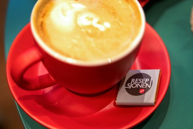 Til venstre: en rød kaffekopp med hjerte tegnet i melka, til høyre: folkemengde inne på Resepsjonen, Kristiansand, under kafeåpning.