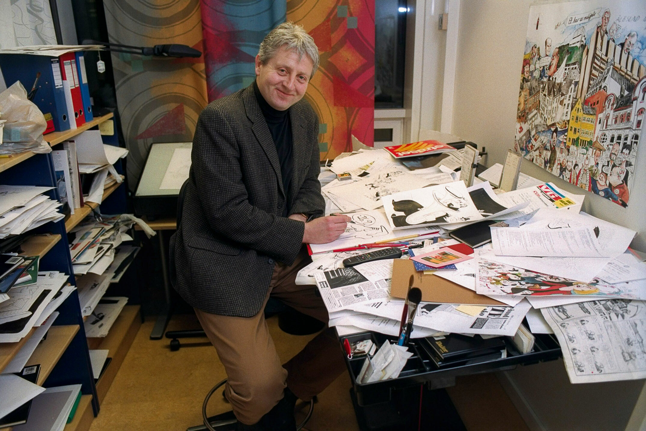 Foto: Mann midt i livet sitter på et kontor med en masse løse ark og tegninger på. Bak ham er det en bokhylle full av ark og permer.