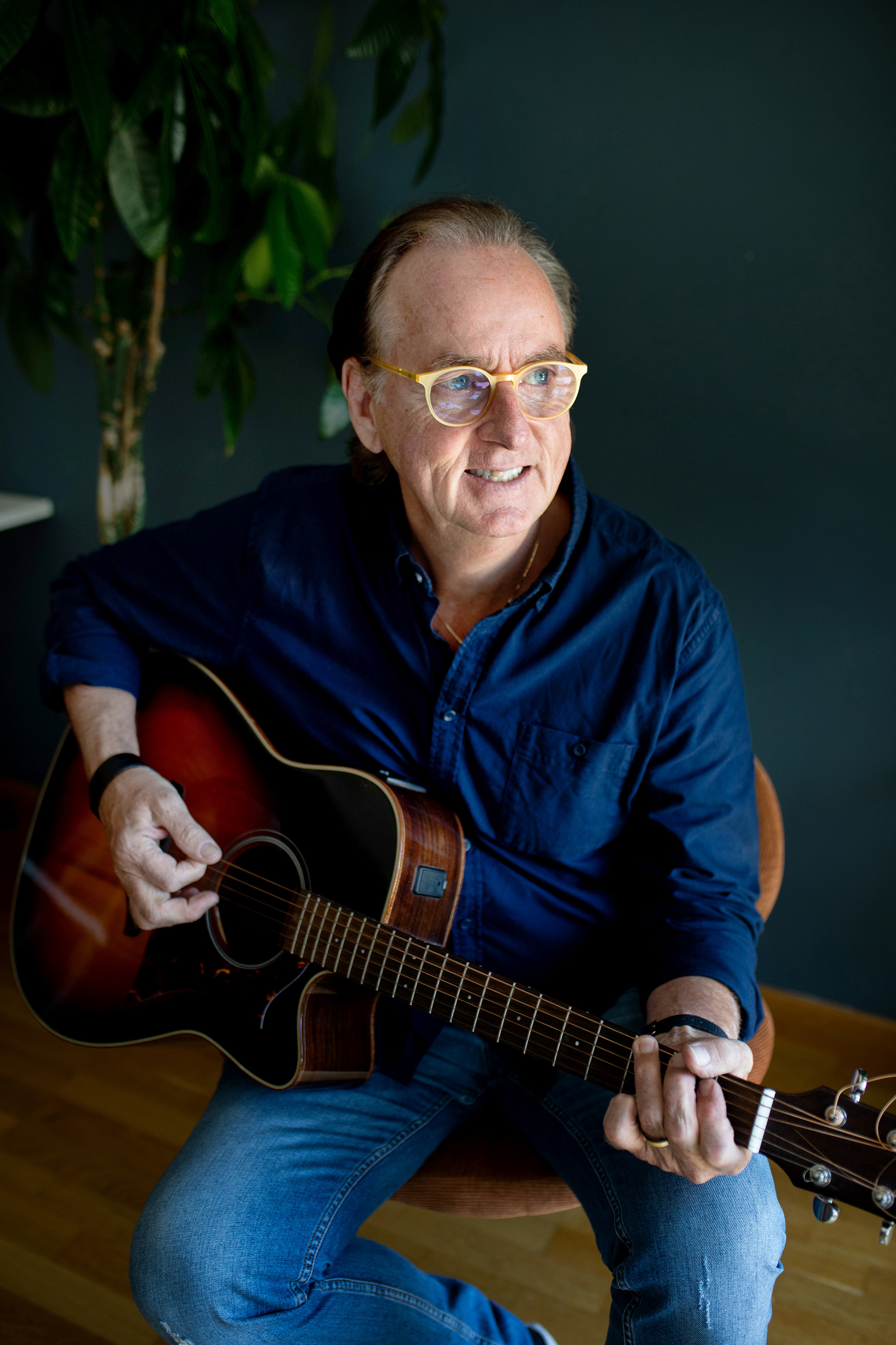 70 år gamle Roland Utbult sitter og spiller på Gitaren. Han har på seg blå skjorte og jeans og smiler inn i kamera