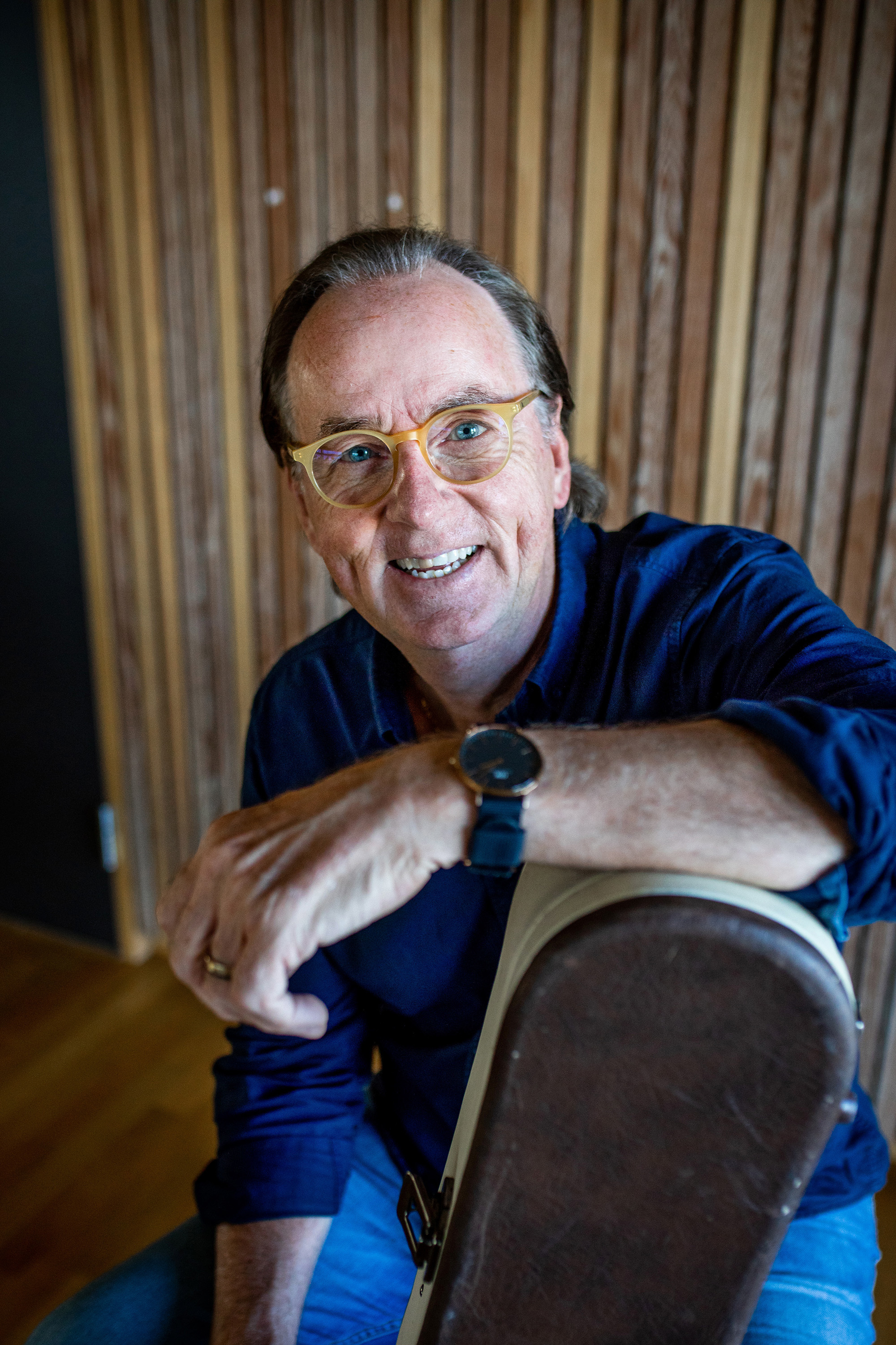 70 år gamle Roland Utbult sitter i blå skjorte og jeans og smiler inn i kamera