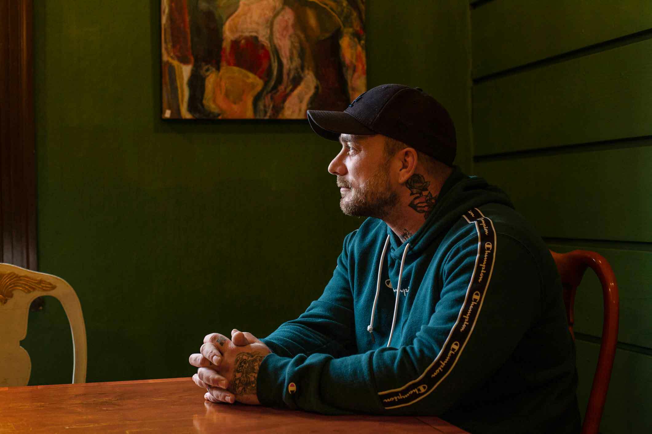 Mann med grønnblå hoodie og kotrtbarbert på hodet sitter på et spisebord med elder stoler. Et kunstverk på den grønne veggen bak. Mannen har skyggelue og tatoveringer på halsen og på fingrene.