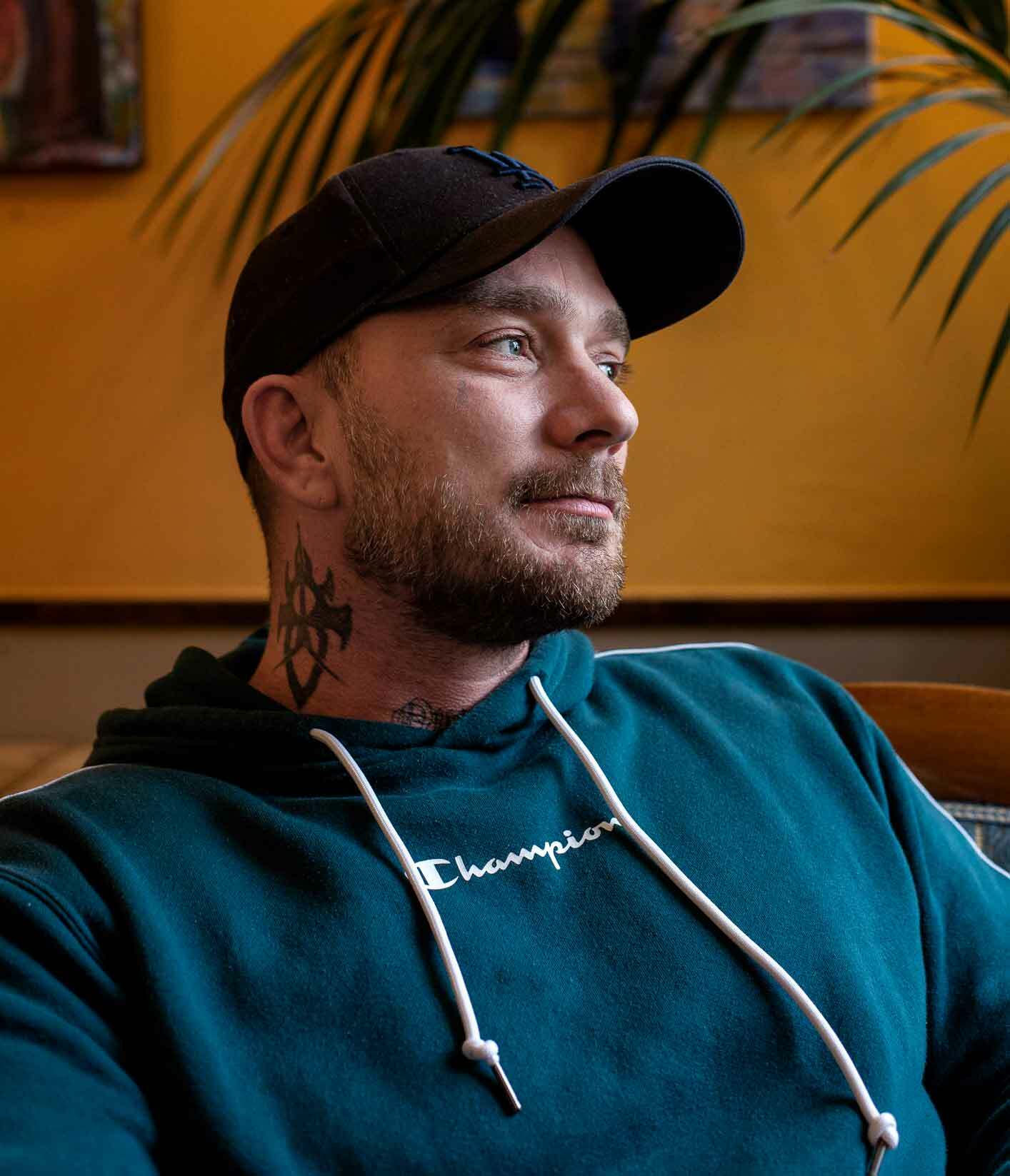 Mann med grønnblå hoodie og kotrtbarbert på hodet. Mannen har skyggelue og tatoveringer på halsen og på fingrene.