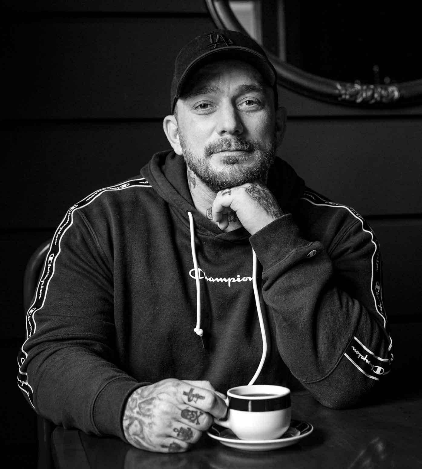 Sort/hvitt-bilde av en mann med hoodie, kortbarbert på hodet og kort skjegg. Han har skyggelue og tatoveringer på halsen og hendene.