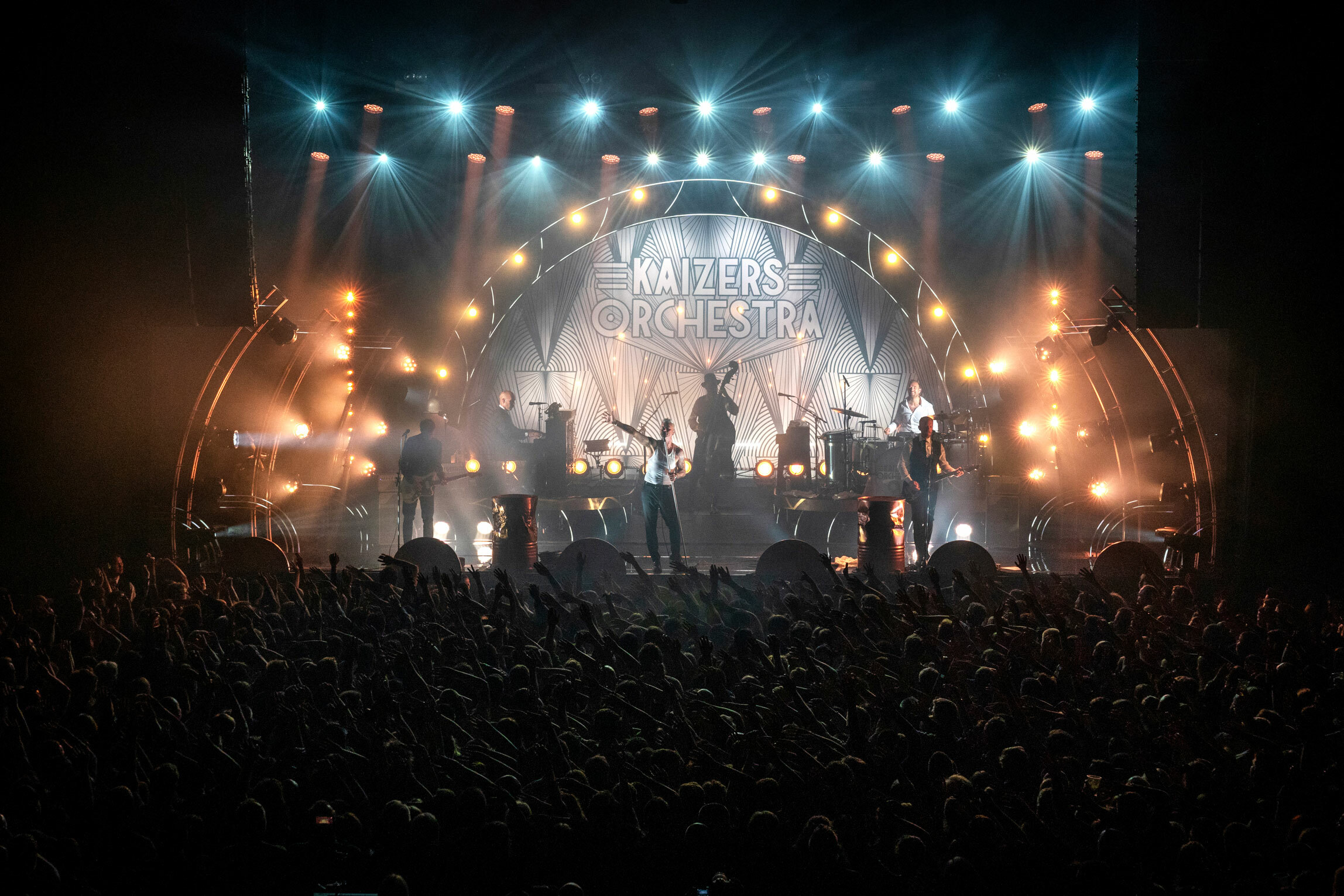 Foto: Stort bilde av hele bandet på scenen. På veggen bak, under alle scenelysene står logoen til Kaizers Orchestra.