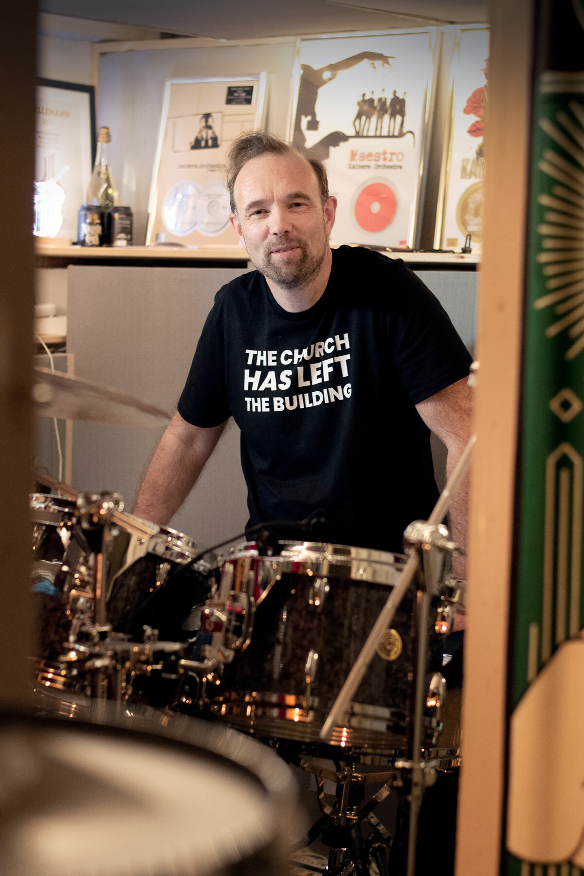 Foto: Mann med mørkt hår og skjegg sees gjennom en døråpning til en kjellerstue. Han sitter ved et slagverk. På hylla over hodet hans står det en rekke tildelte priser.