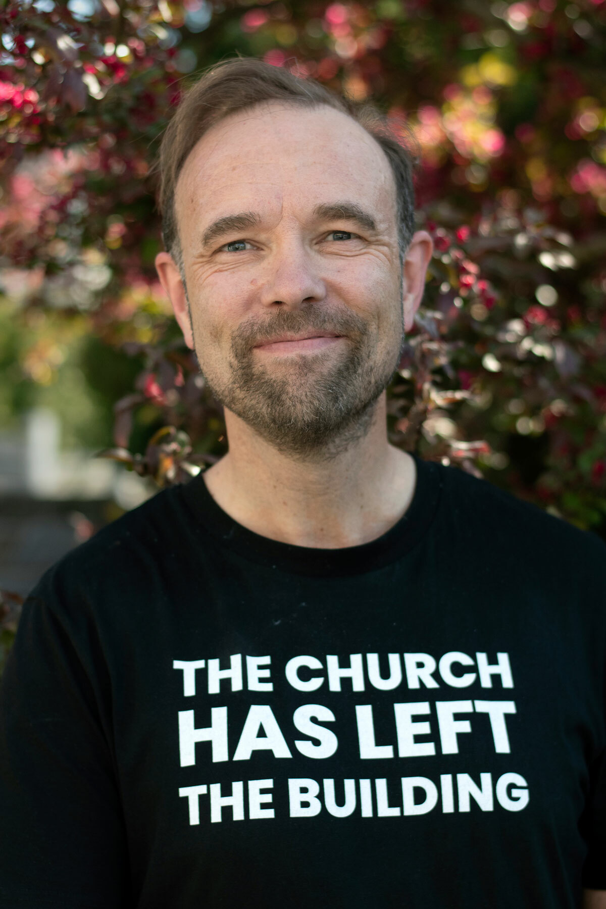 Foto: Mann med mørkt hår og skjegg. Han har på seg en svart T-skjorte med hvit skrift der det står: The church has left the building.