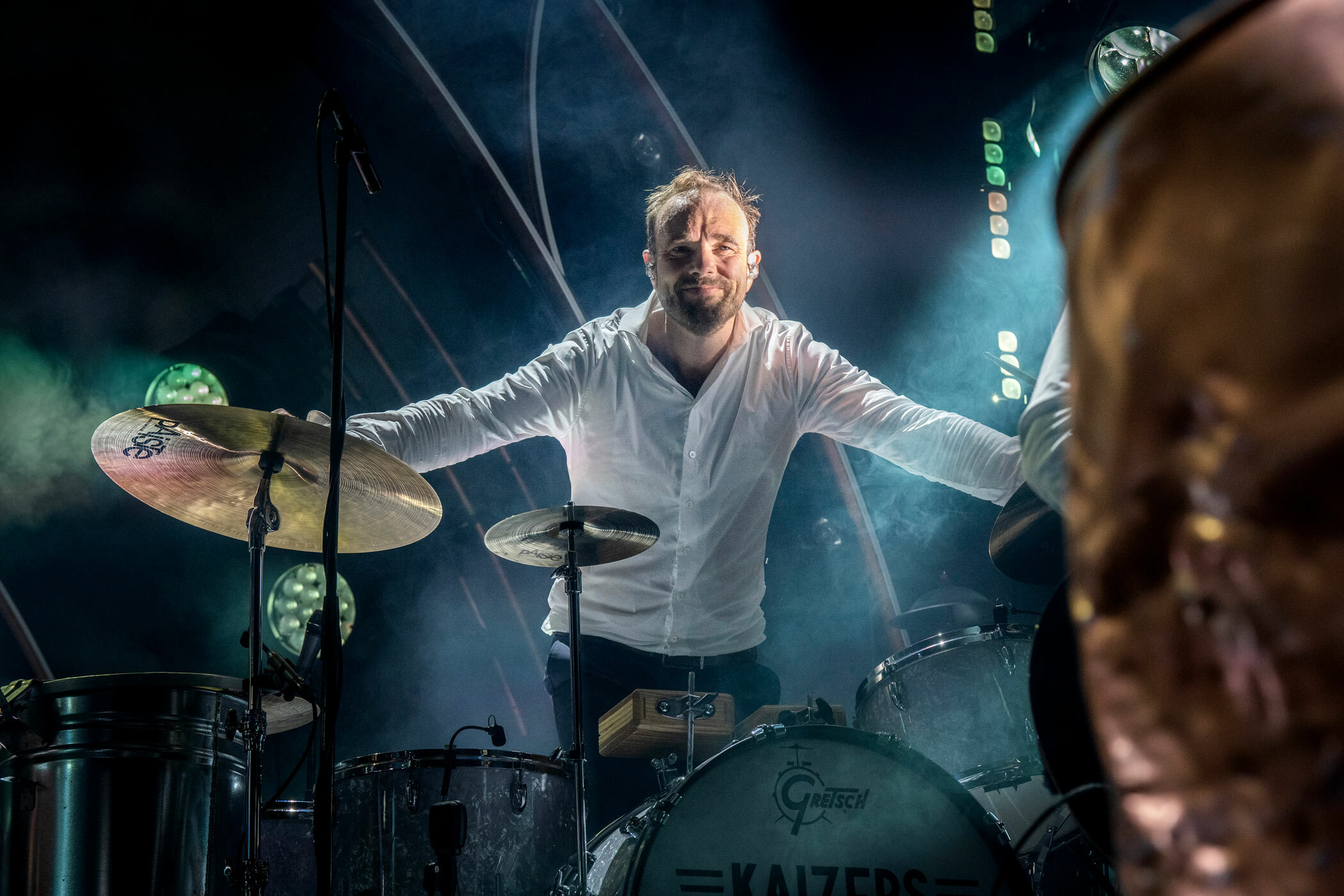 Foto: Mann med hår og mørkt skjegg smiler svett bak et trommesett. I hvit skjorte står han svett med spotlighten rett i ansiktet.