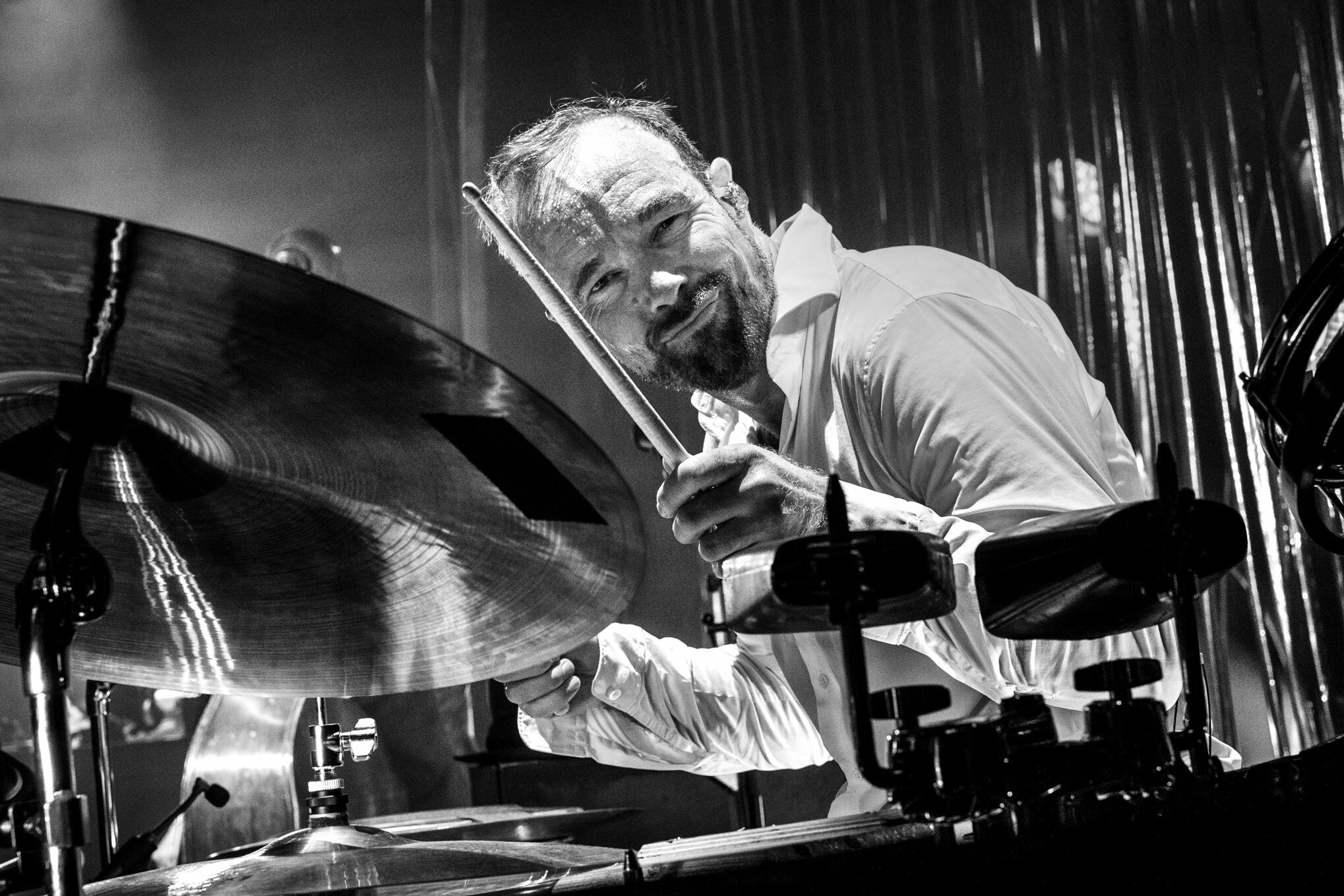Foto: Svart-hvitt-bilde av trommeslager med mørkt hår og skjegg som lener seg inn mot den ene symbalen mens han spiller. Ser rett i kamera.