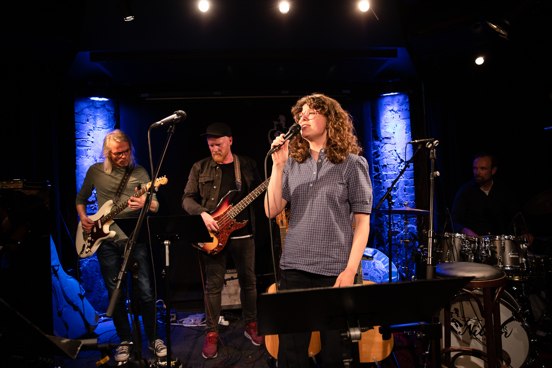Dame med briller og mørke krøller som synger. I bakgrunnen står bandet.