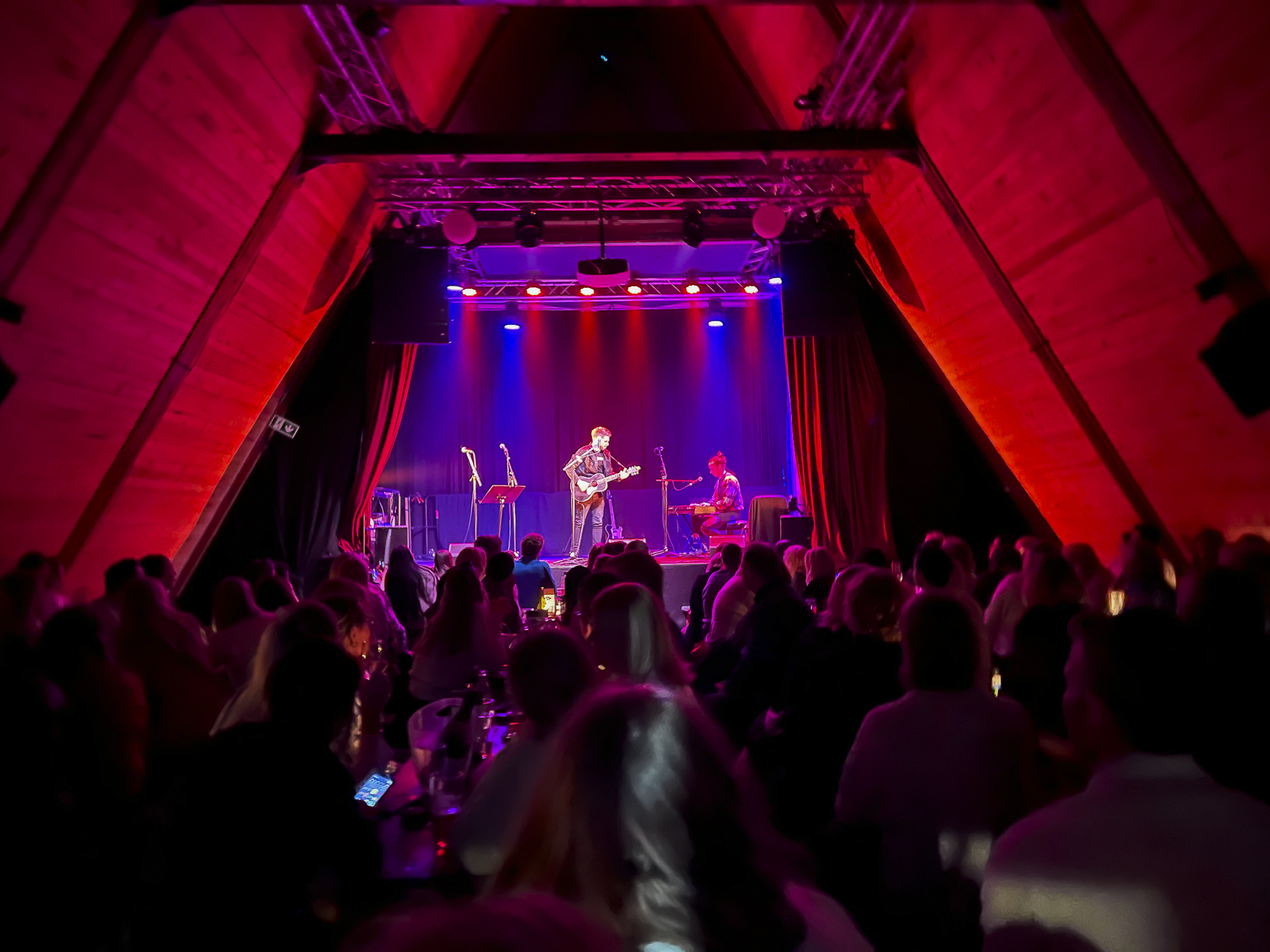 En artist står på en scene  som er opplyst av rødt og blålilla lys. Lokalet har spisst skråtak og publikum ser vi bakfra.