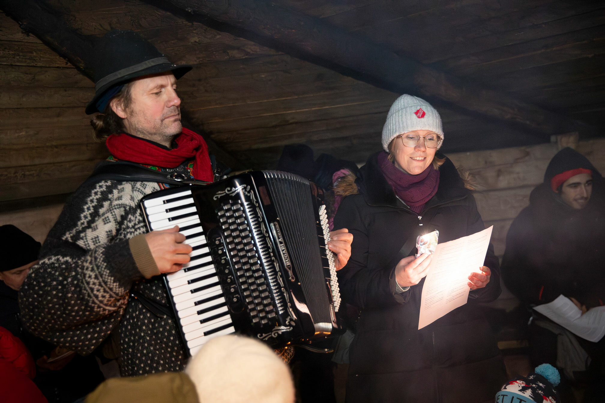 Foto: Mann i strikkengenser, rødt skjerf og hatt spiller trekkspill. Ved siden av ham sitter en dame med mobil og lyser opp et ark med sangtekster med mobilen sin. Det sitter flere folk rundt i en lavvo i tre.