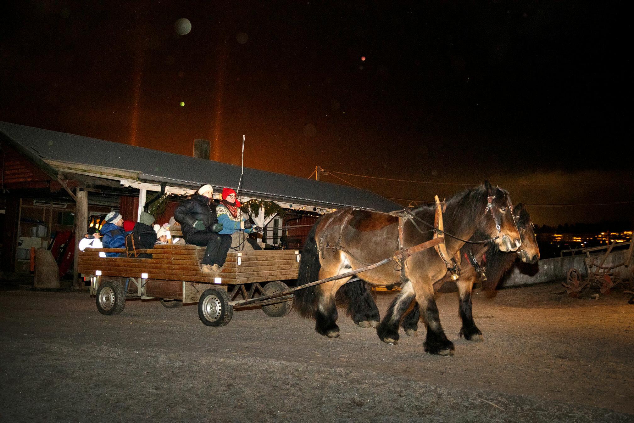 Foto: To brune hester drar en kjerre full av barn og voksne gjennom mørket. Det er litt snø på bakken og en gårdsbygning i bakgrunnen.