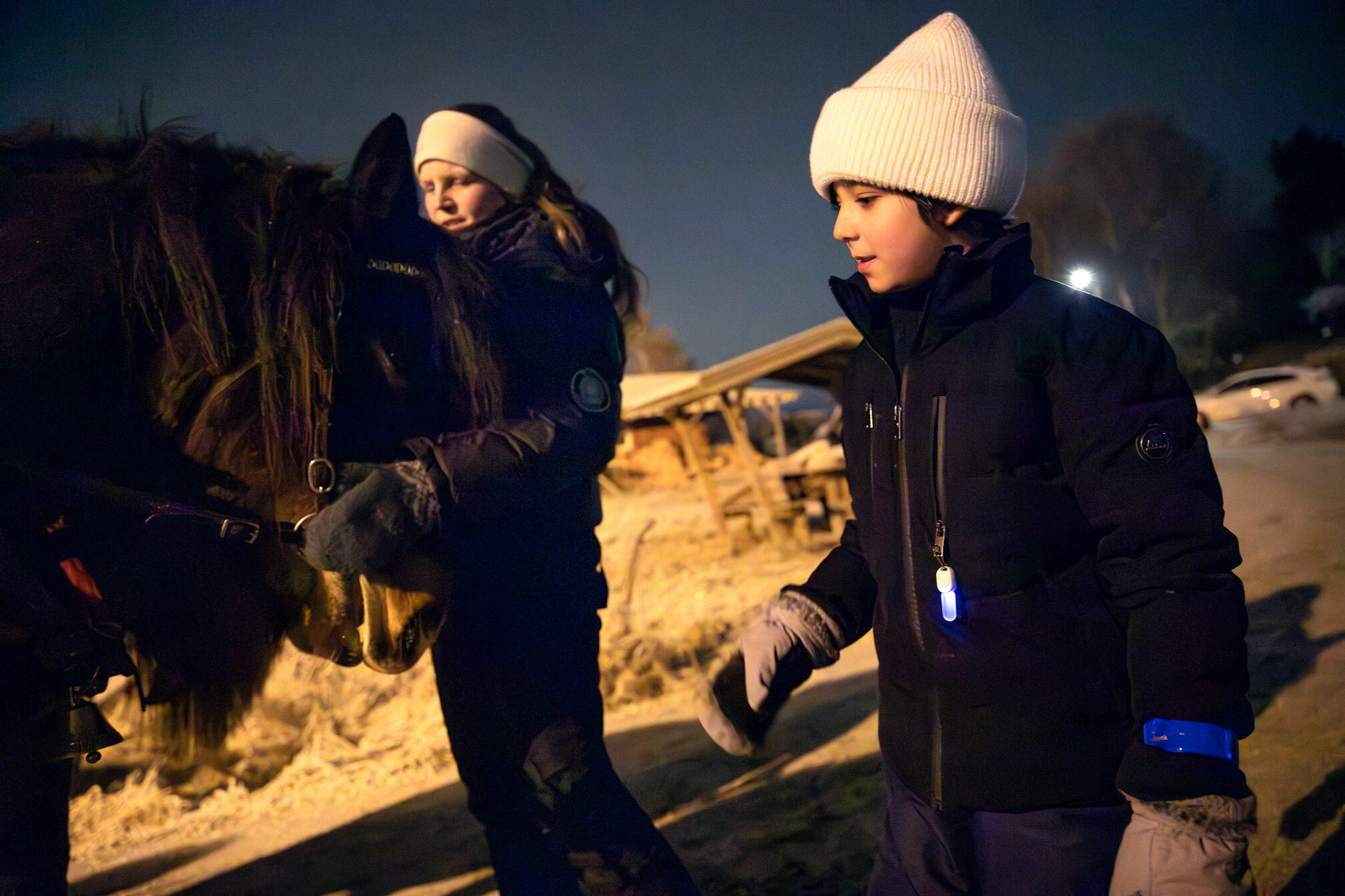 Foto: Kvinne med langt, blondt hår og hvitt pannebånd klapper en av hestene mens en gutt rundt åtte år med mørkt hår under den hvite topplua går forbi hestene. Ute er det kaldt så her gjelder det å ha på seg boblejakke og hansker.