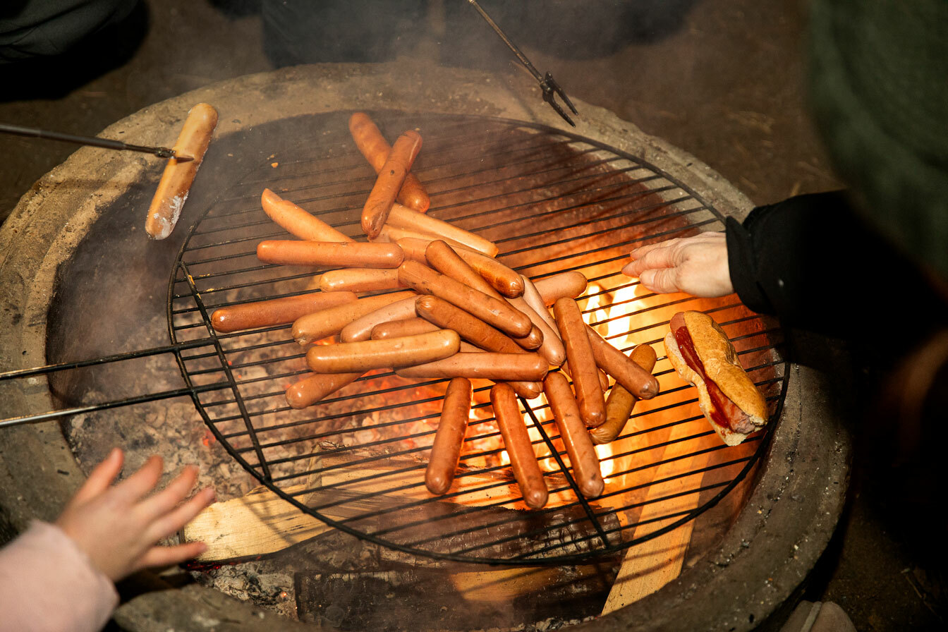 Foto: Pølser som grilles over åpen flamme og barn som varmer hender der.