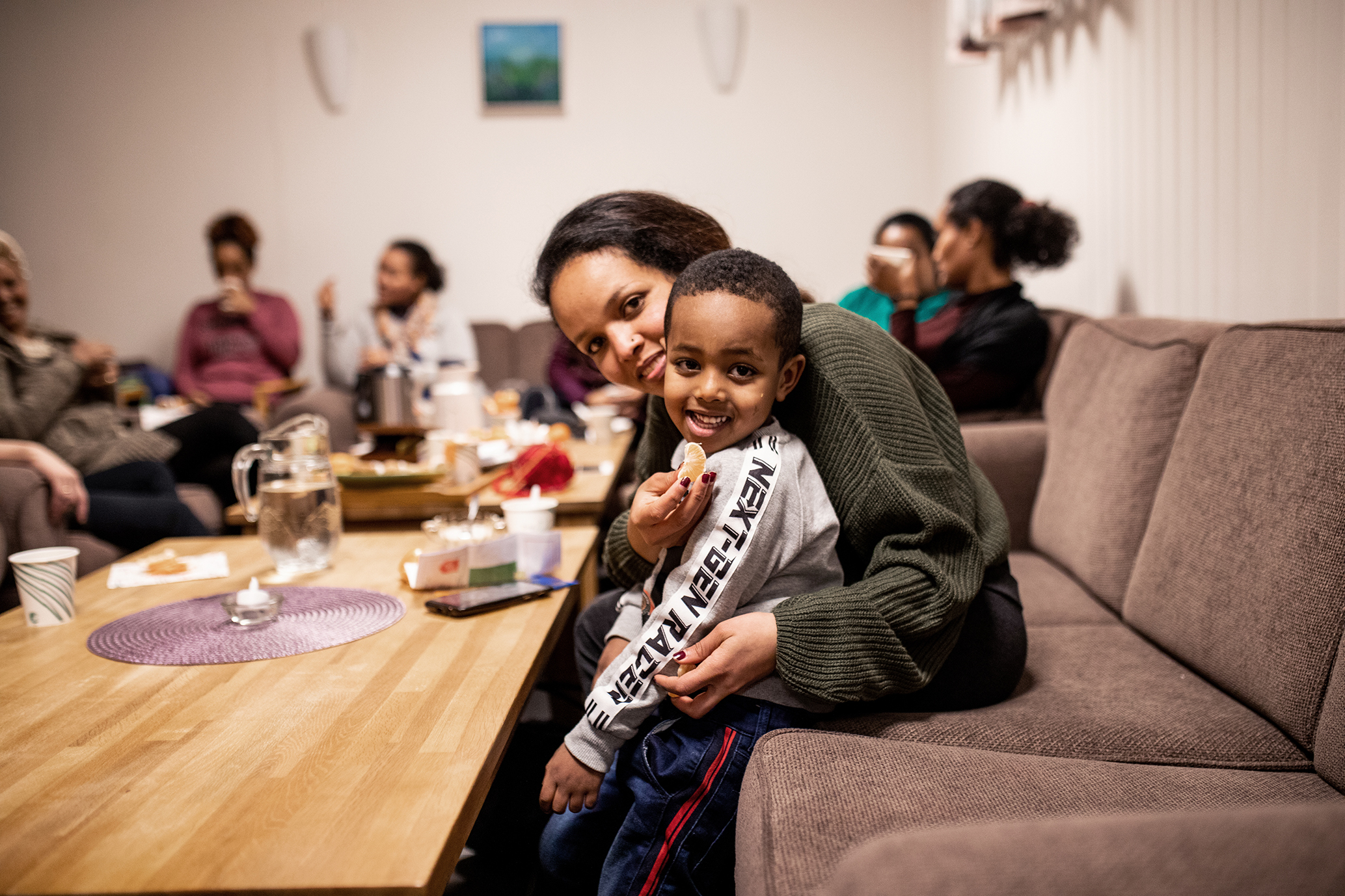 Bildet viser en kvinne som sitter i en sofa og holder rundt en liten gutt. I bakgrunnen kan vi skimte mange andre kvinner som sitter og prater.