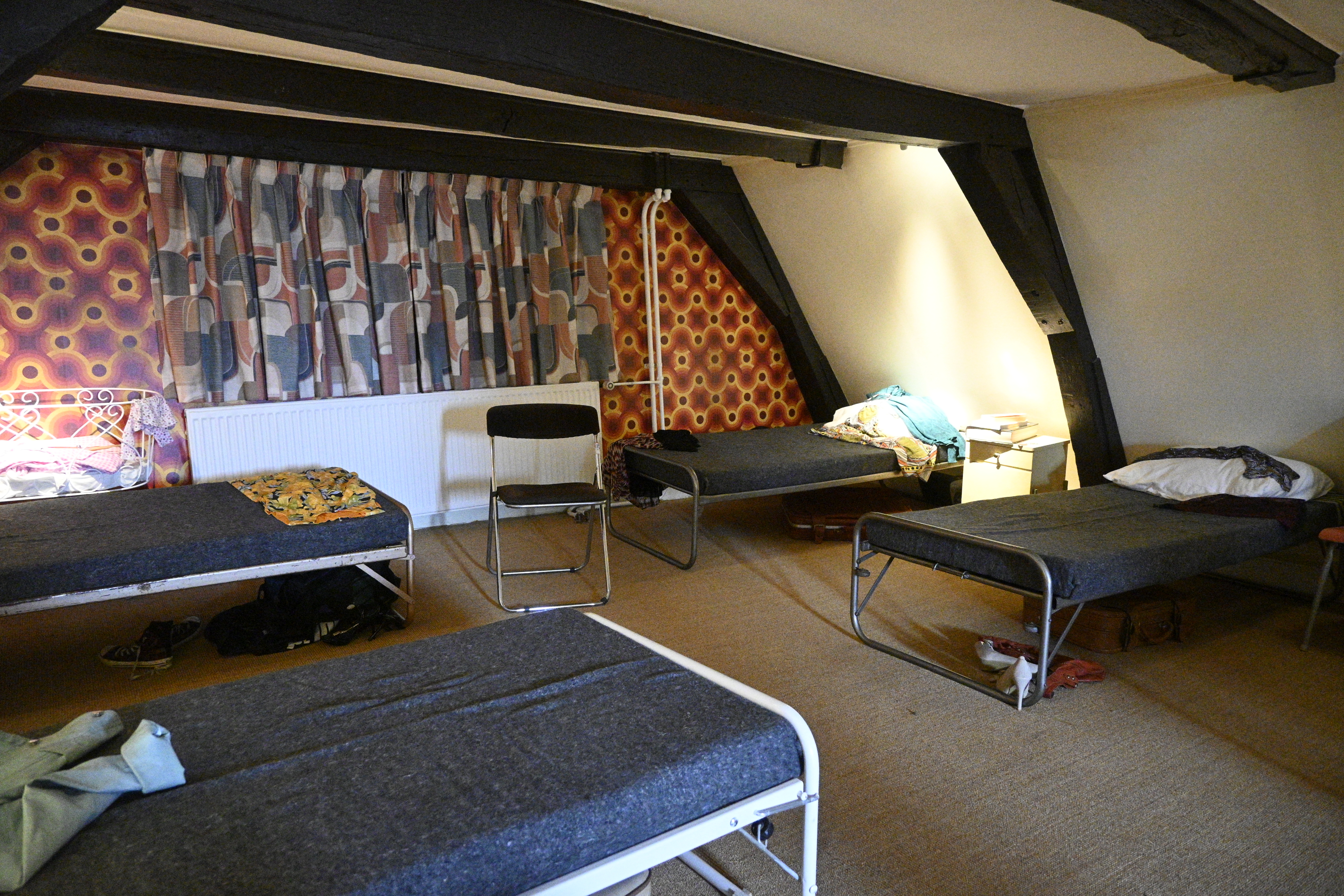 Et loftsrom med fire feltsenger langs veggene. Det er 70-tallstapet og -tekstiler på endeveggen og bjelker i taket