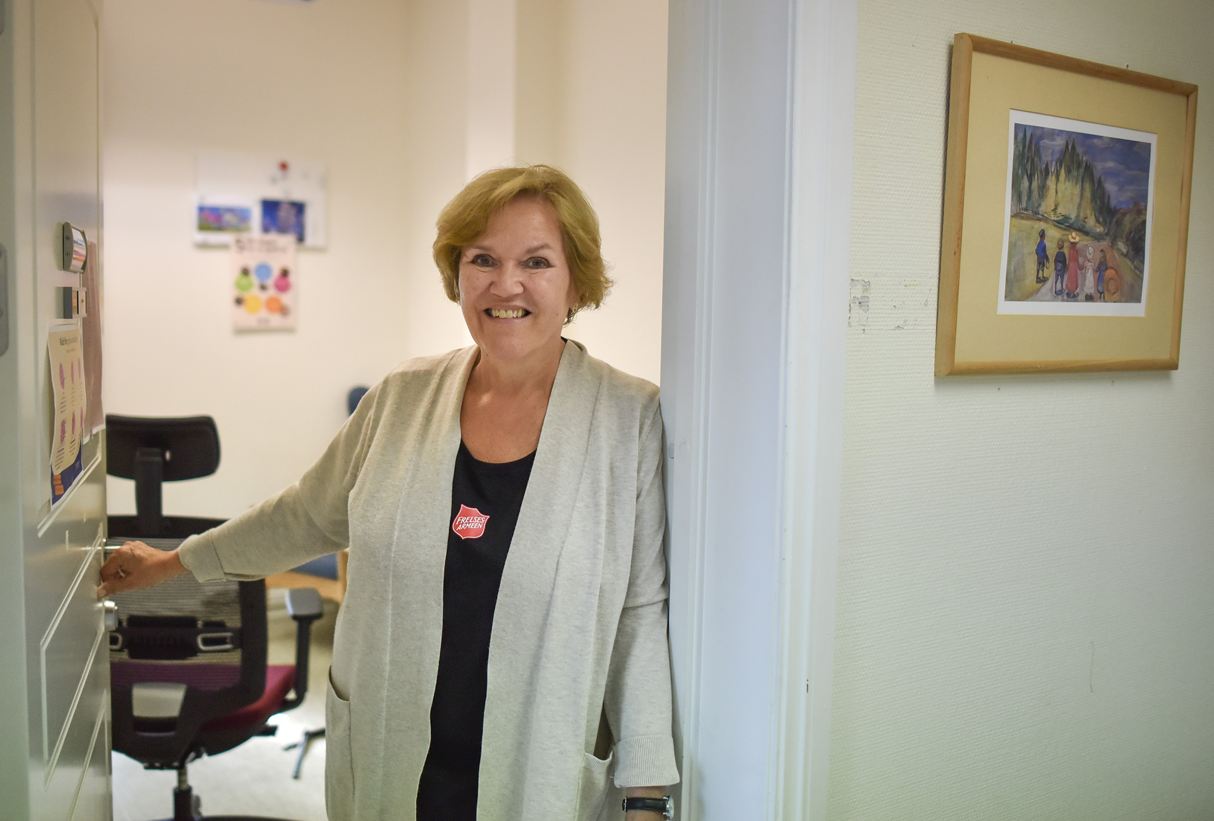 Foto: Kvinne med mørkt hår i 50-årene står i en døråpning inn til et kontor. Hun har på seg svart T-skjorte med logo fra Frelsesarmeen og en beige strikkejakke.