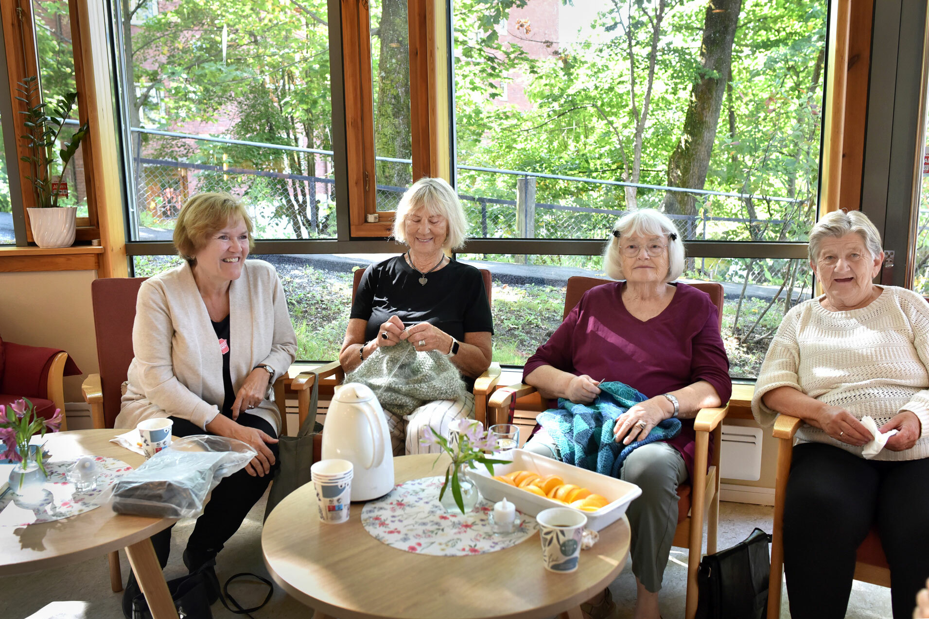 Foto: Fire kvinner sitter på rekke langs et vindu, den den ene er i 50-årene og de andre tre i 70-årene. De to i midten sitter med hvert sitt strikketøy.