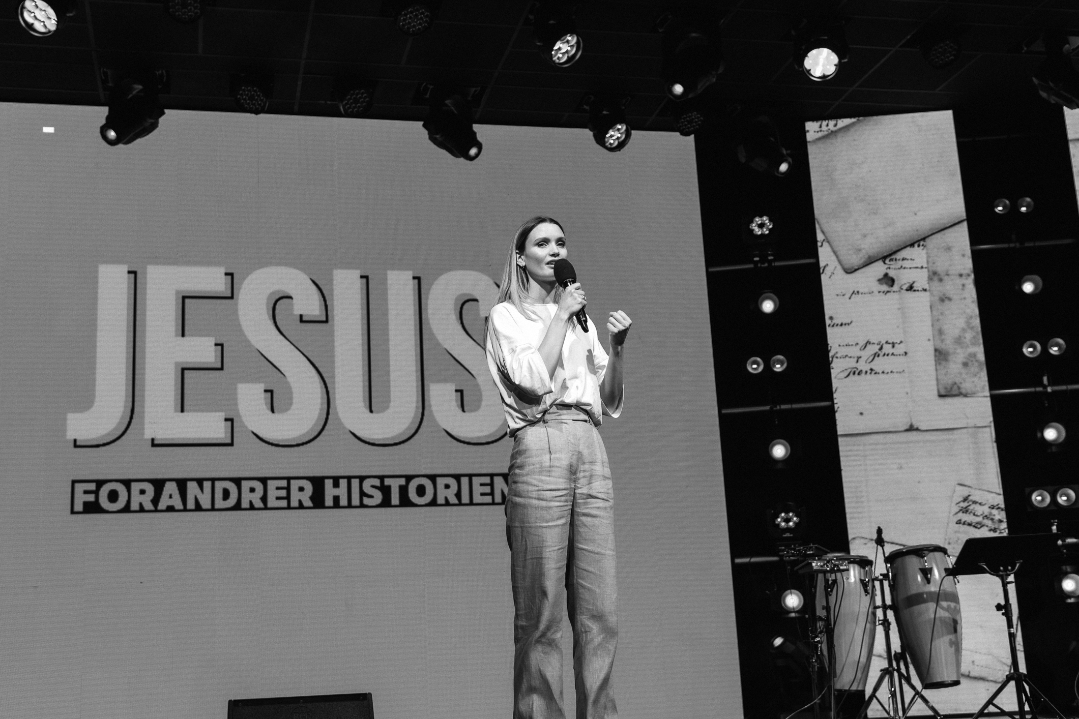 Foto: I svart/hvitt av kvinne foran på en scene. Hun snakker i mikrofon. På skjermen bak står det i store bokstaver: "Jesus forandrer historien".
