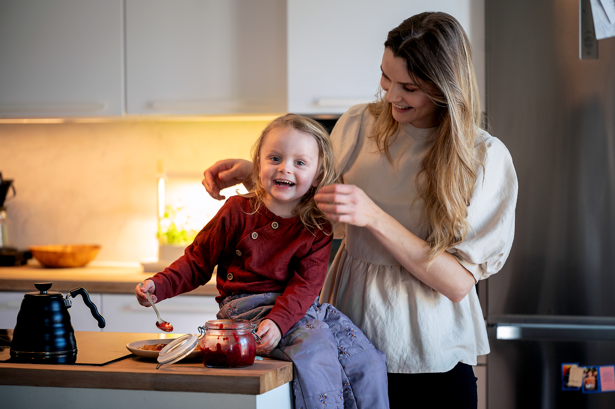 To foto av mor og datter på kjøkkenet. Treåringen ser inn i kamera og ler på begge bildene. Moren ser på datteren.