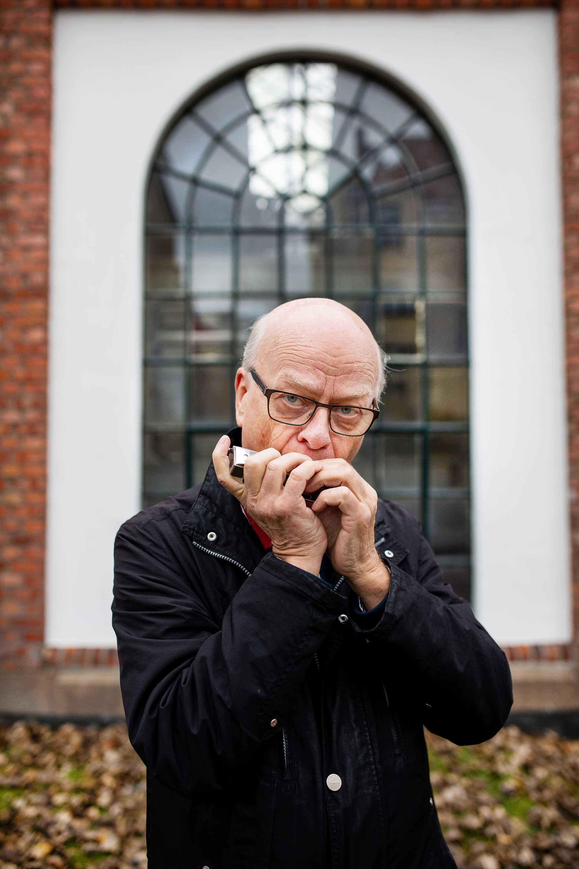 Sigmund Groven står ute, foran ett stort buet vindu, og spiller på munnspillet.