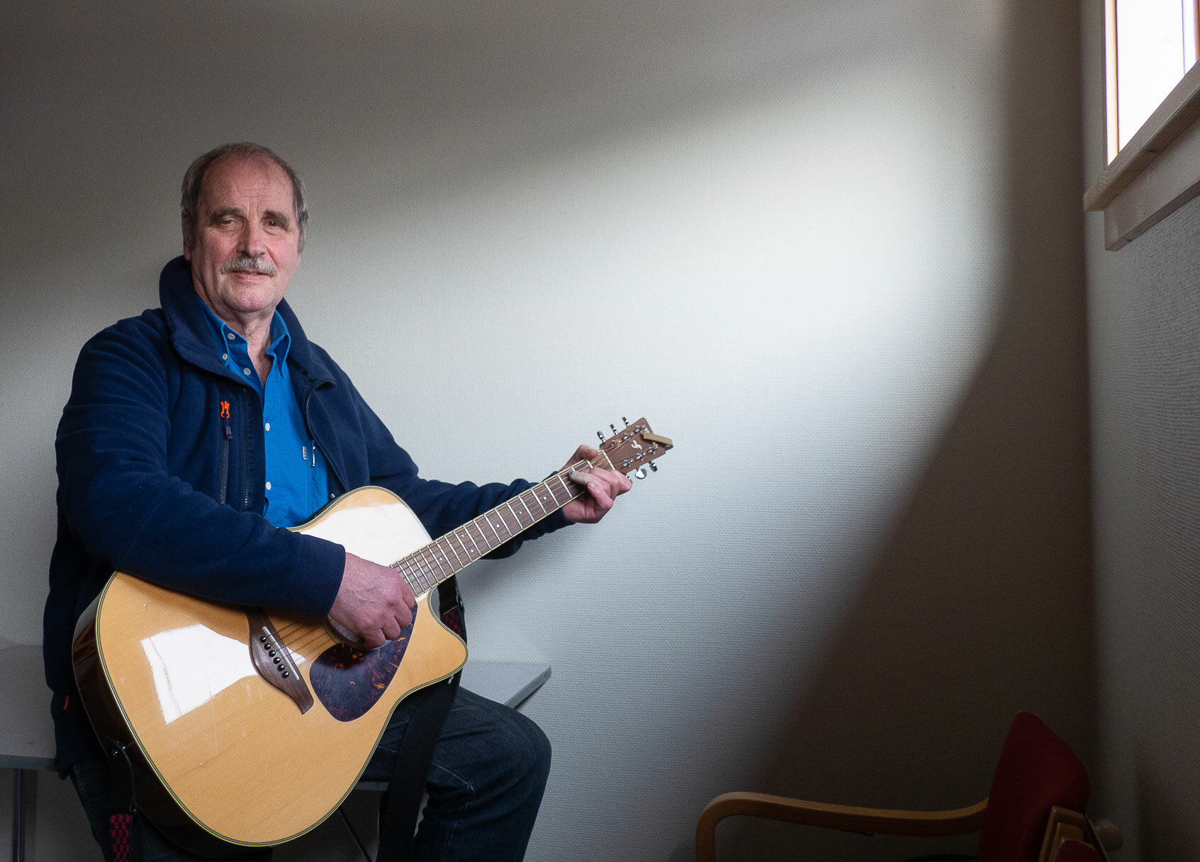 Mann med gitar sitter i et litt mørkt rom. Lyset fra et lite vindu oppe på veggen stråler ned på ham.
