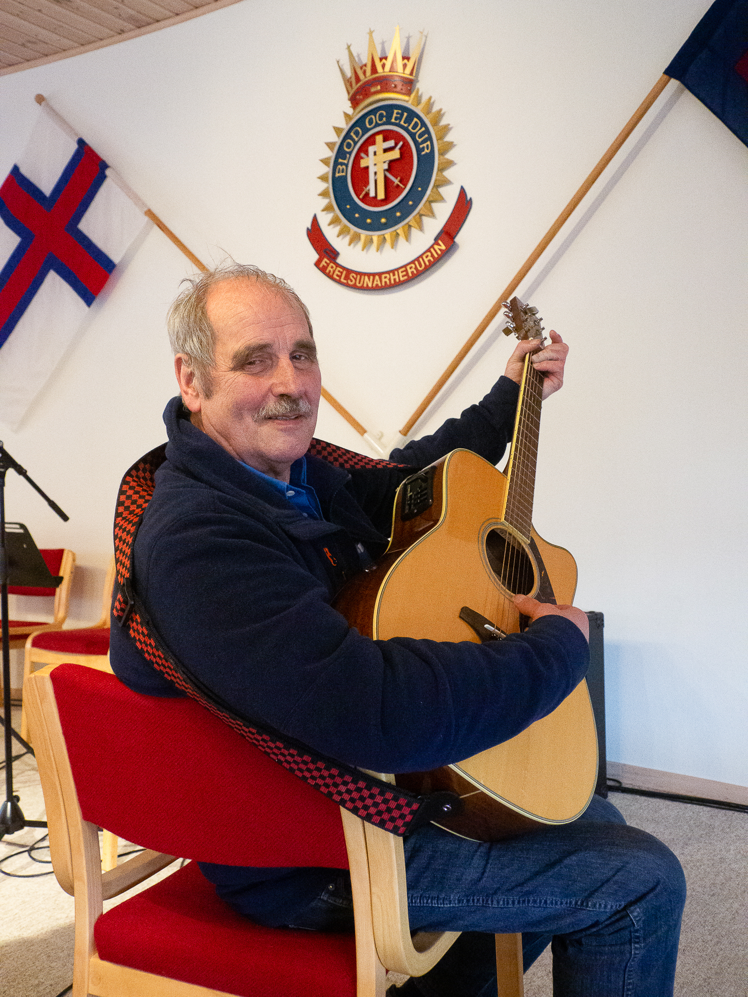 Mann med gitaren inne i et rom med færøysk flagg på veggen sammen med Frelsesarmeens krone, som er et slags våpenskjold.