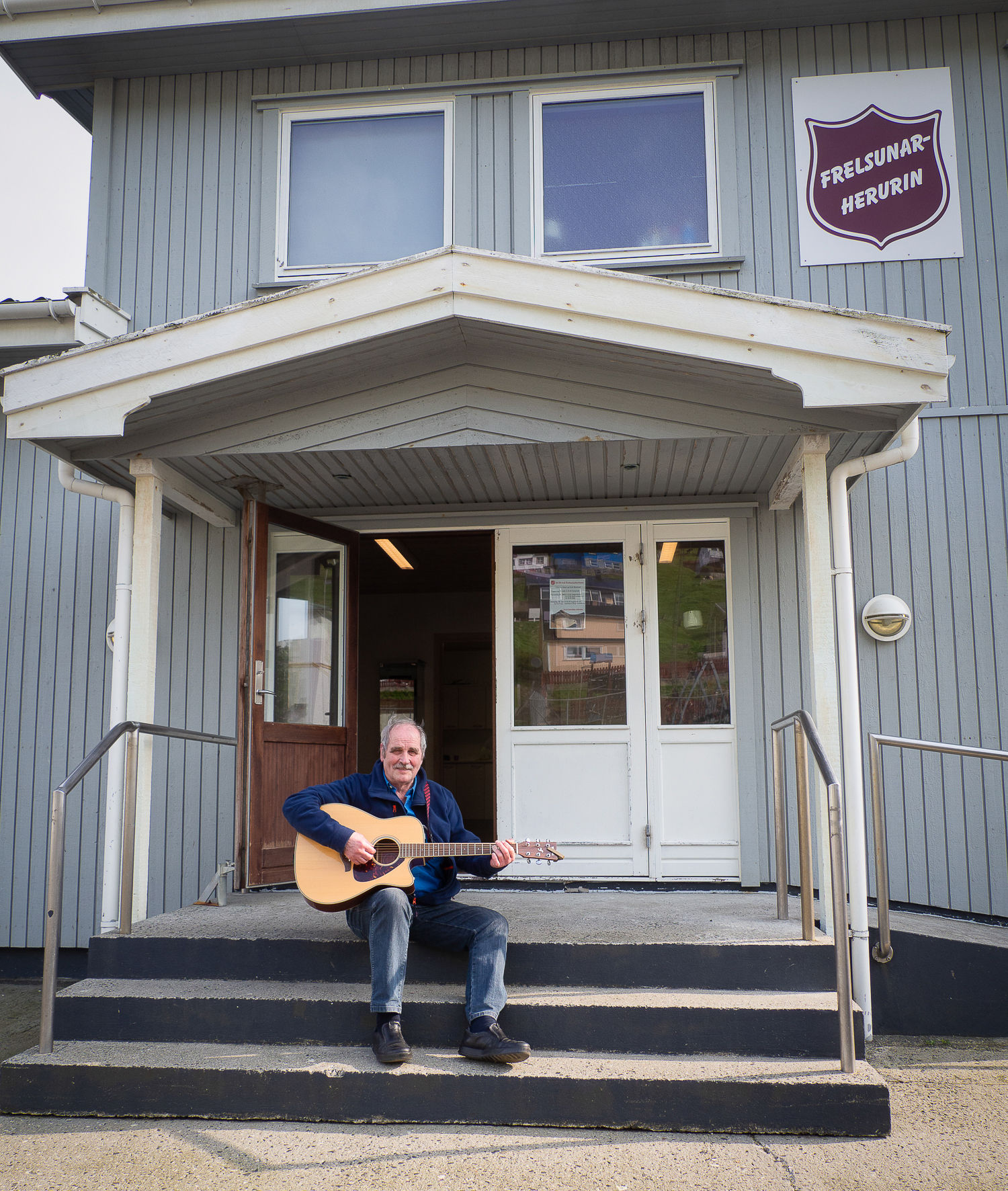 Mann sitter på trappa til et grått trehus med Frelsesarmeens logo på. Han spiller gitar.