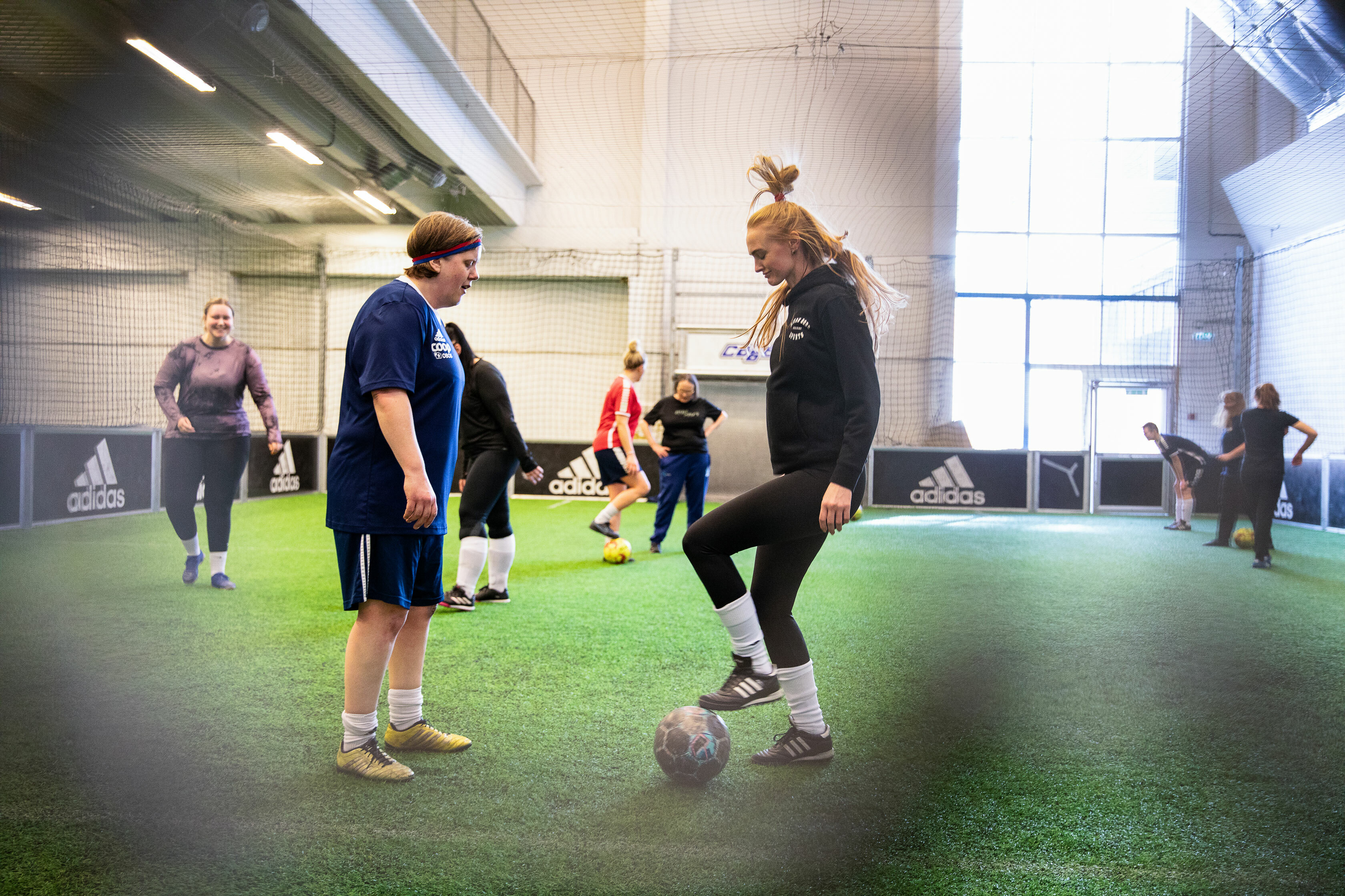 Foto: To damer som sparker fotball innendørs på en grønn bane. Flere andre kvinnelige spillere i bakgrunnen.