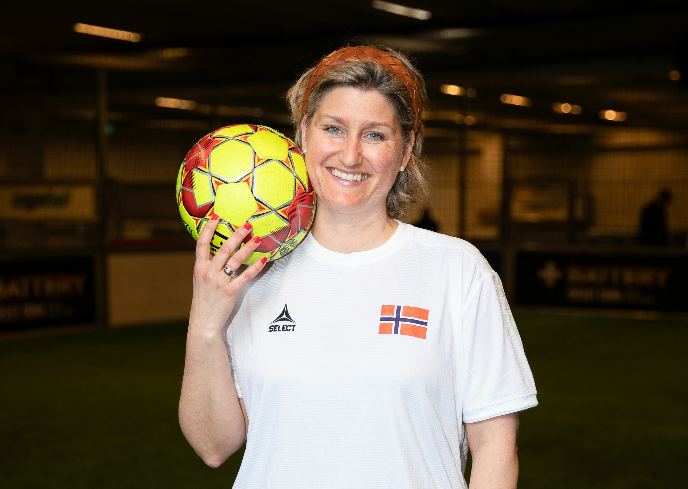 Foto: Kvinne i hvit T-skjorte med et lite, norsk flagg på brystet holder en fargerik fotball ved skulderen. Smiler i kamera.