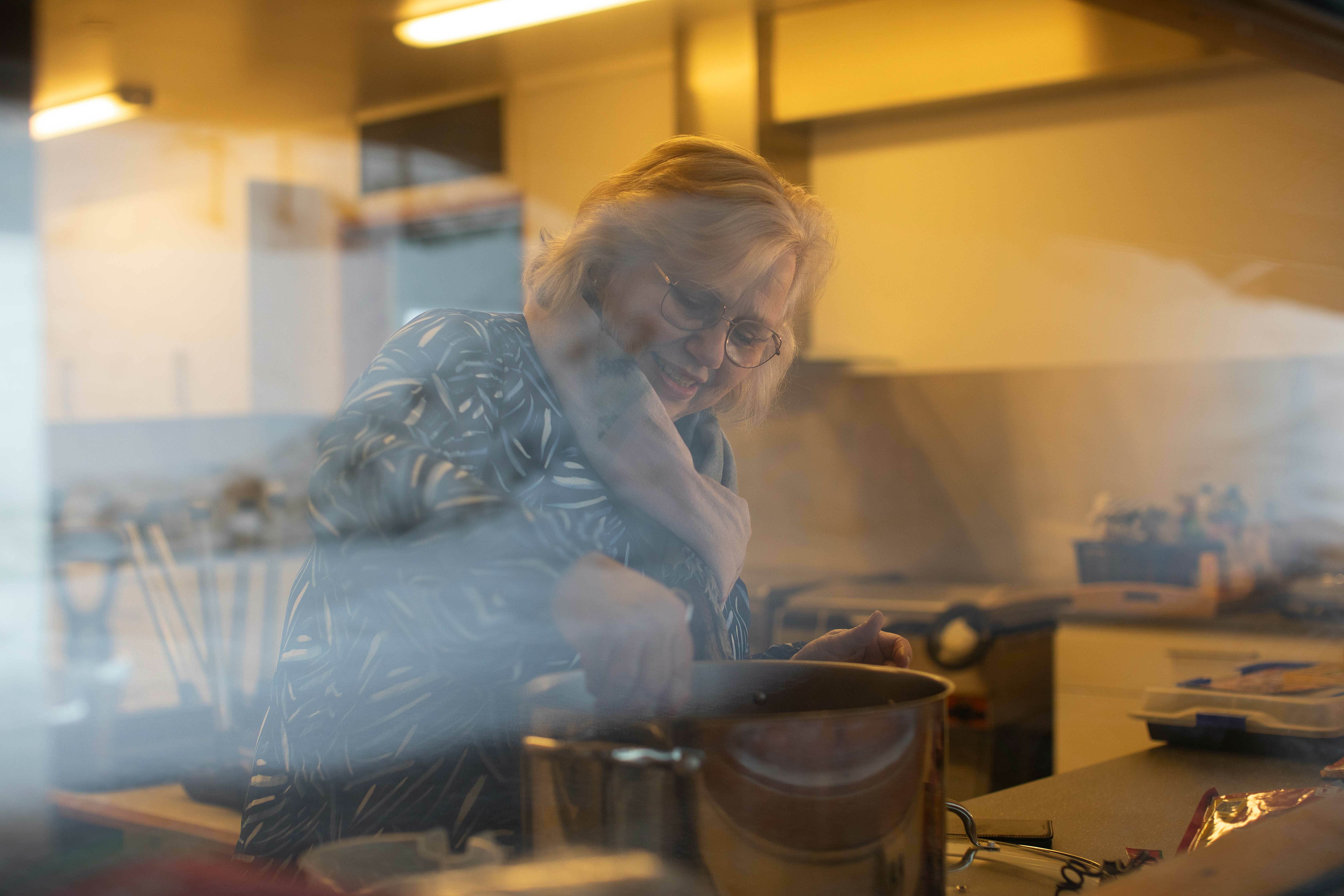 Gjennom en rute ser vi den samme damen koke mat på kjøkkenet.