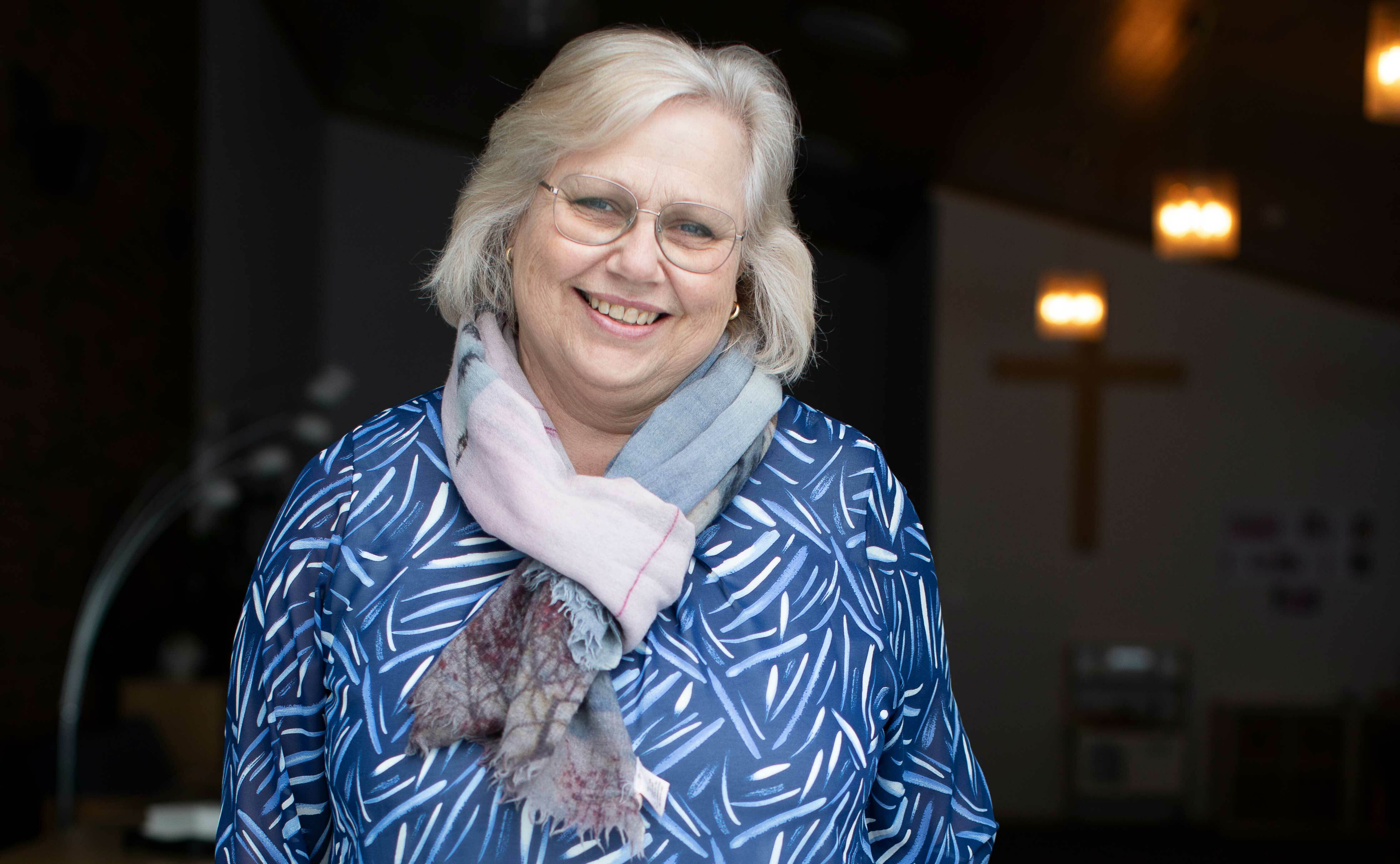 Blond dame med briller smiler mot kamera. Hun er midt i 50-årene. Bak henne i rommet skimter man et kors.