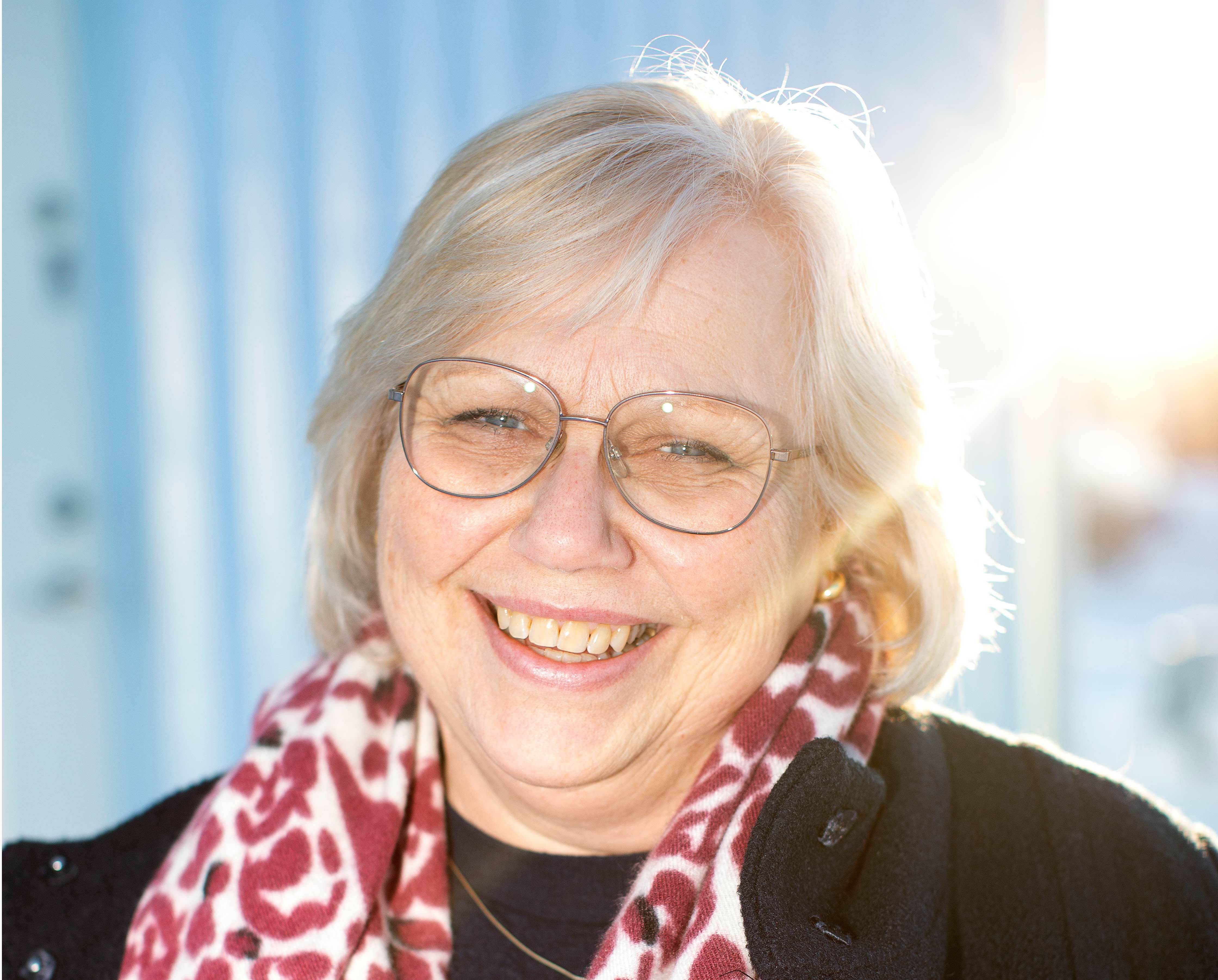 Samme dame med briller smiler. Står utendørs og solen skinner bak henne, gjennom det blonde håret.