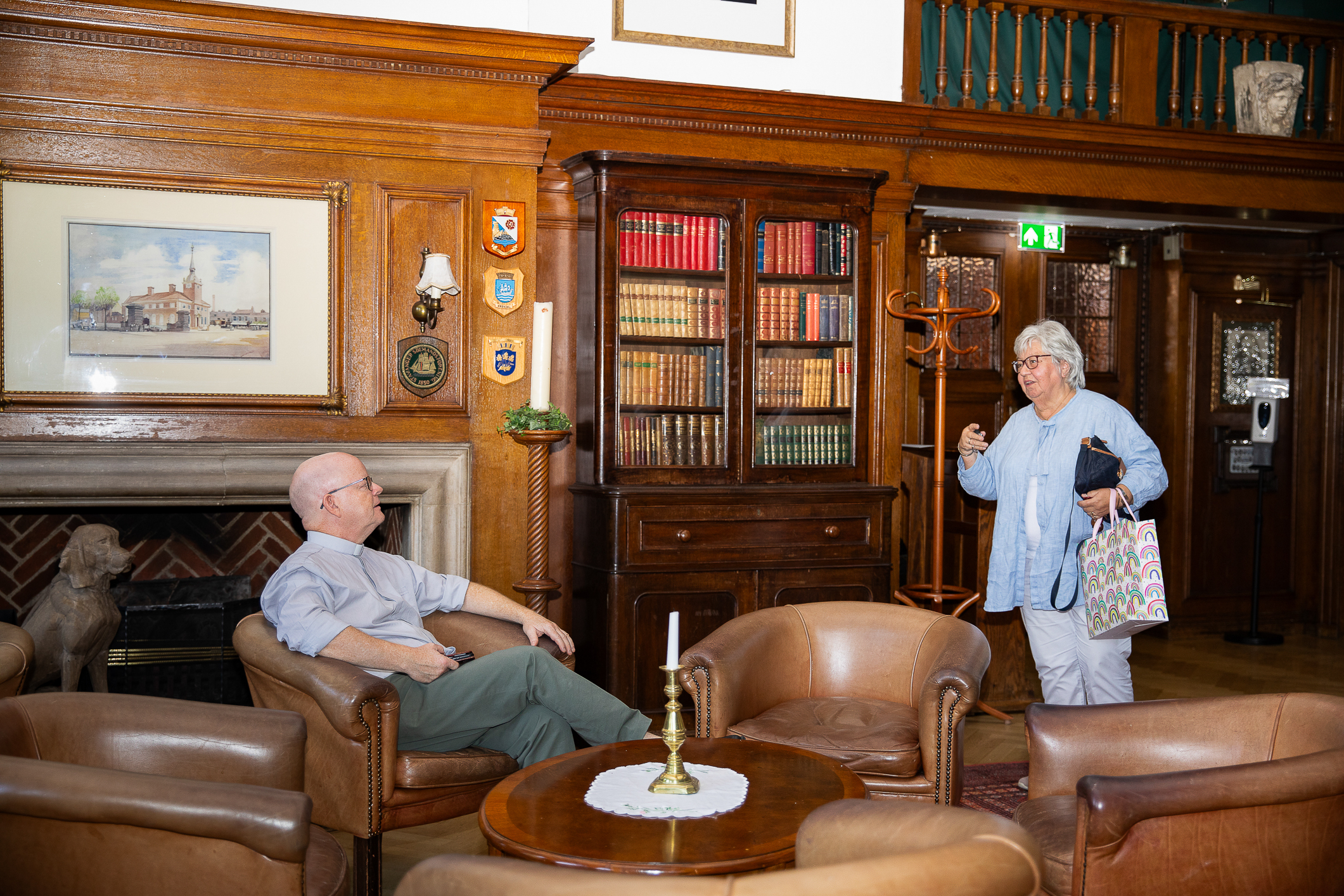 Sofagruppe i skinn i en peisestue. Mann sitter i en stol og kvinne står og snakker med ham. Bøker i skap bak henne.