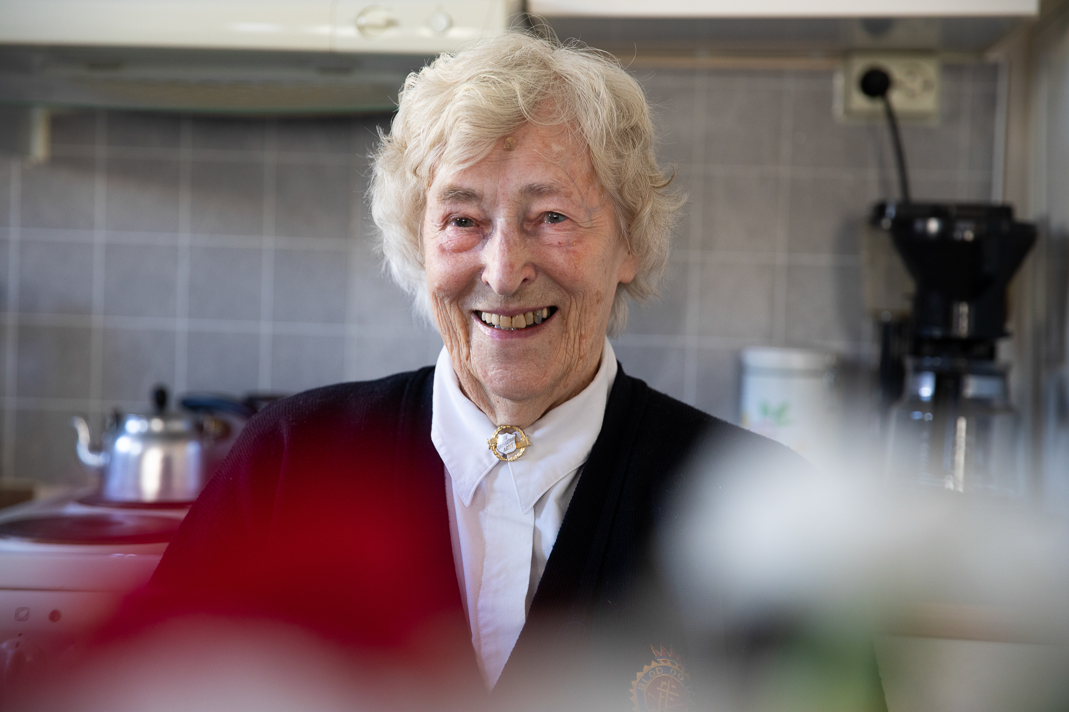 Eldre kvinne med hvitt hår sitter i kjøkkenet sitt og smiler. Hun har hvit bluse og mørk frelsesarméjakke med en slumsøster-nål tett i halsen.