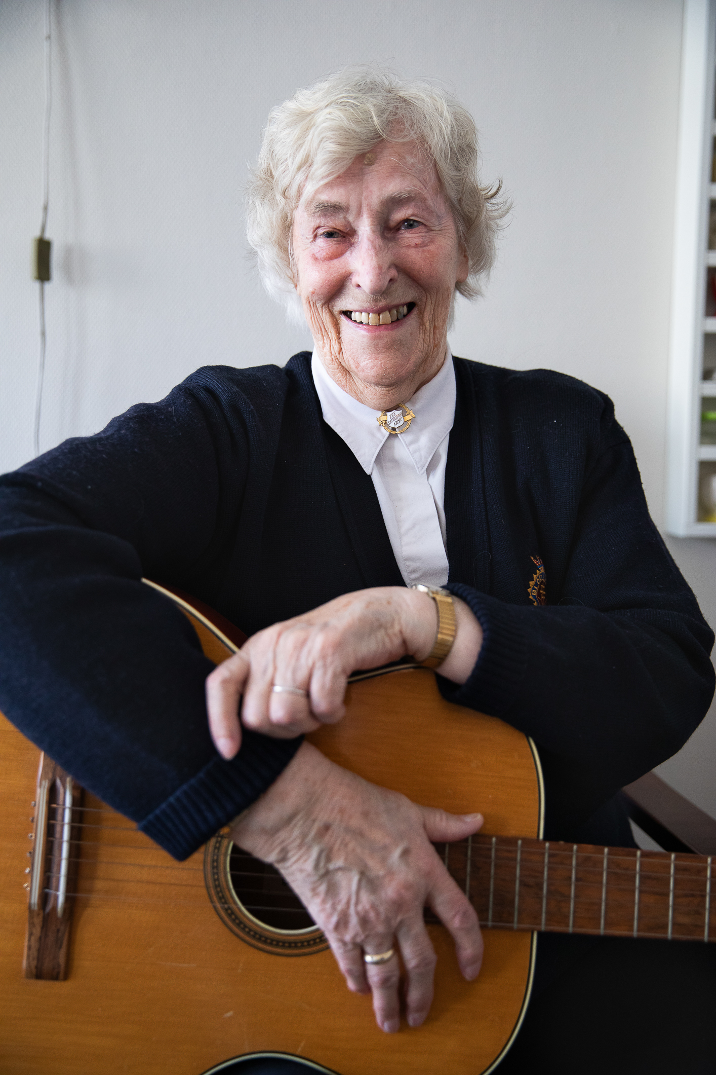 Eldre kvinne med hvitt hår, skjorte med en slumsøster-nål tett i halsen. Mørk frelsesarméjakke.